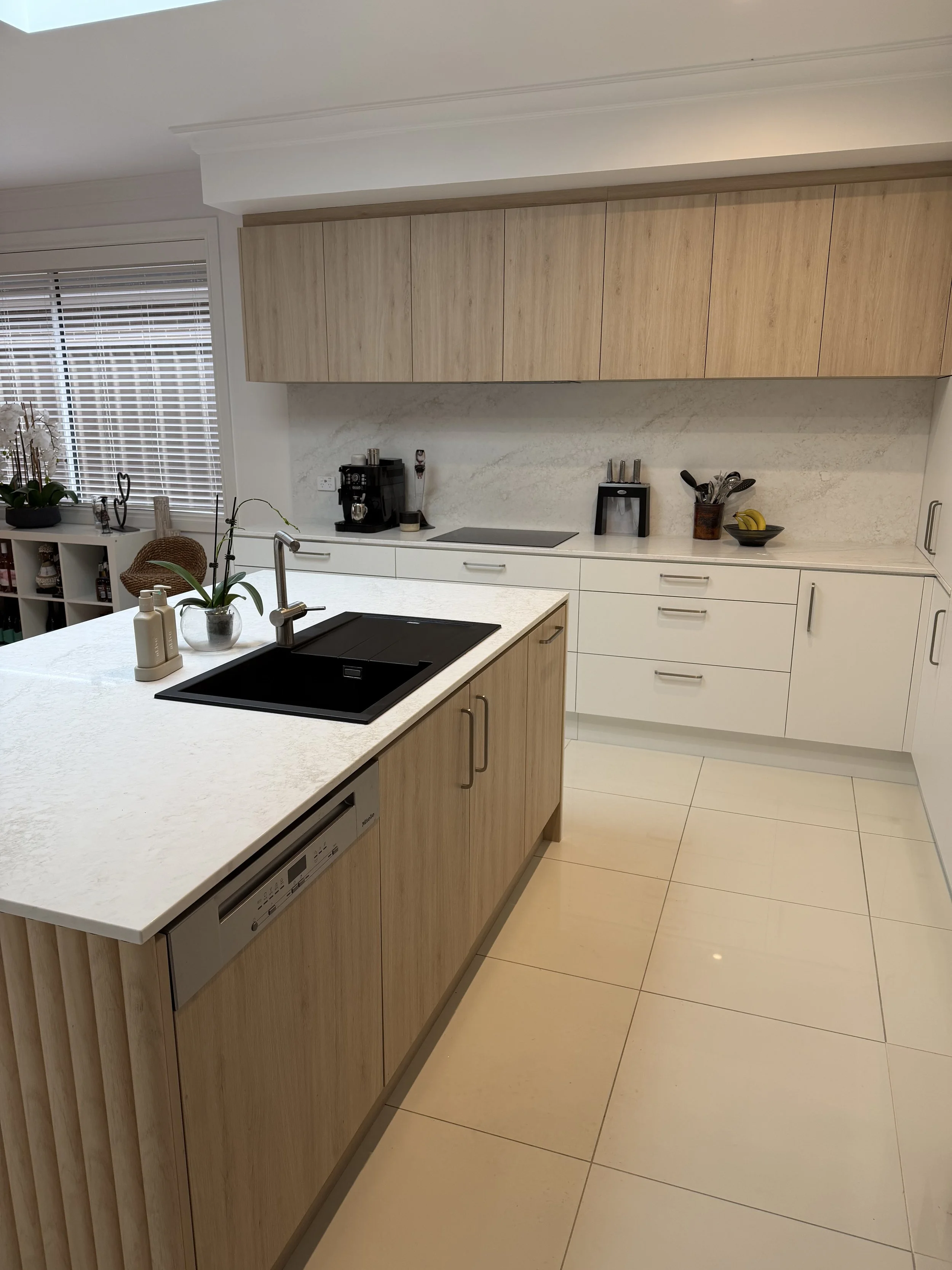 Modern kitchen with white cabinetry, a kitchen island with a black sink, light wood upper and lower cabinets, a window with blinds, a coffee machine, a bowl of bananas, and various kitchen utensils.