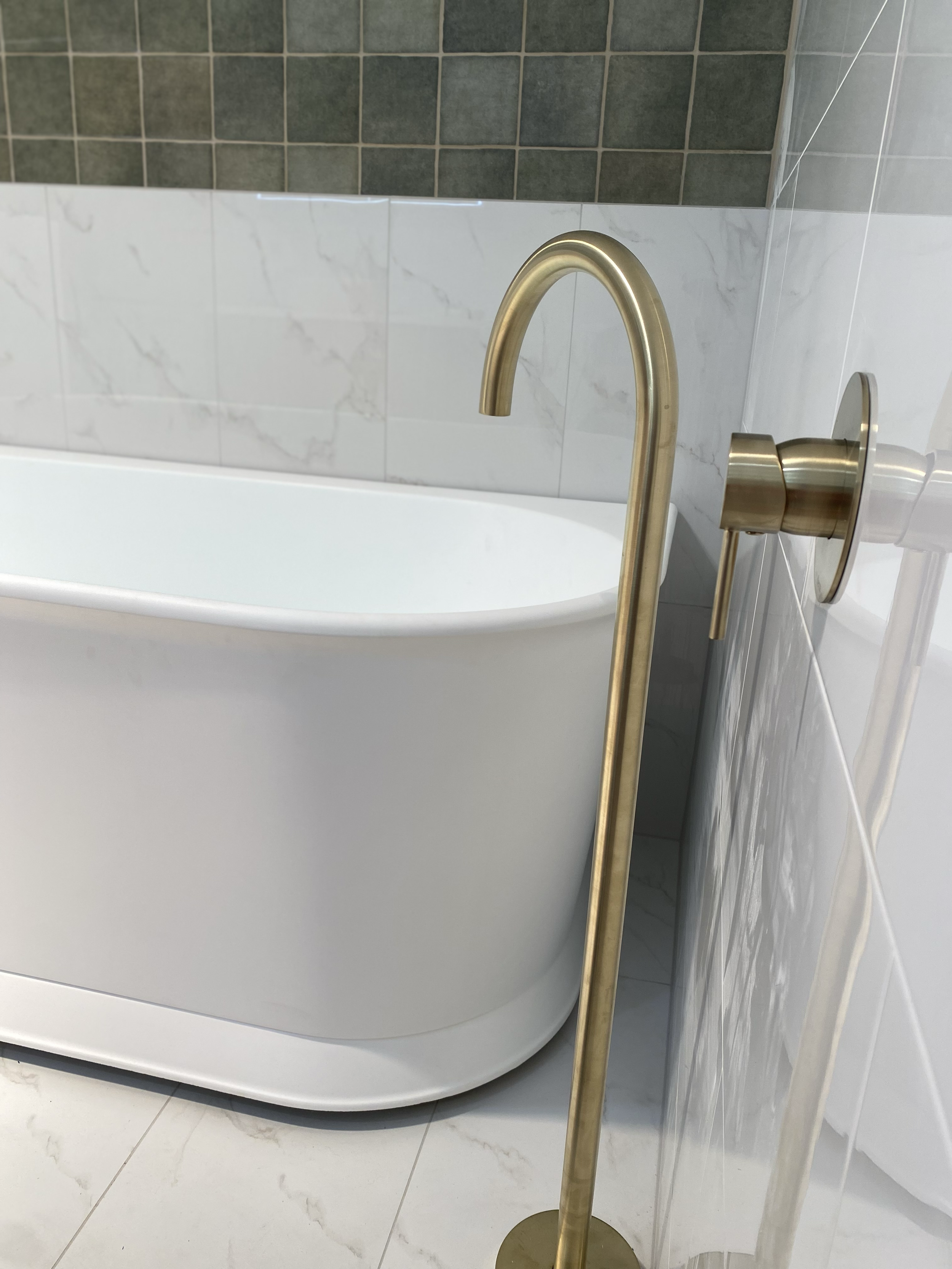 Close-up of a modern bathtub with a gold-colored floor-mounted faucet in a bathroom with white and dark green tiled walls and white marble floor tiles.