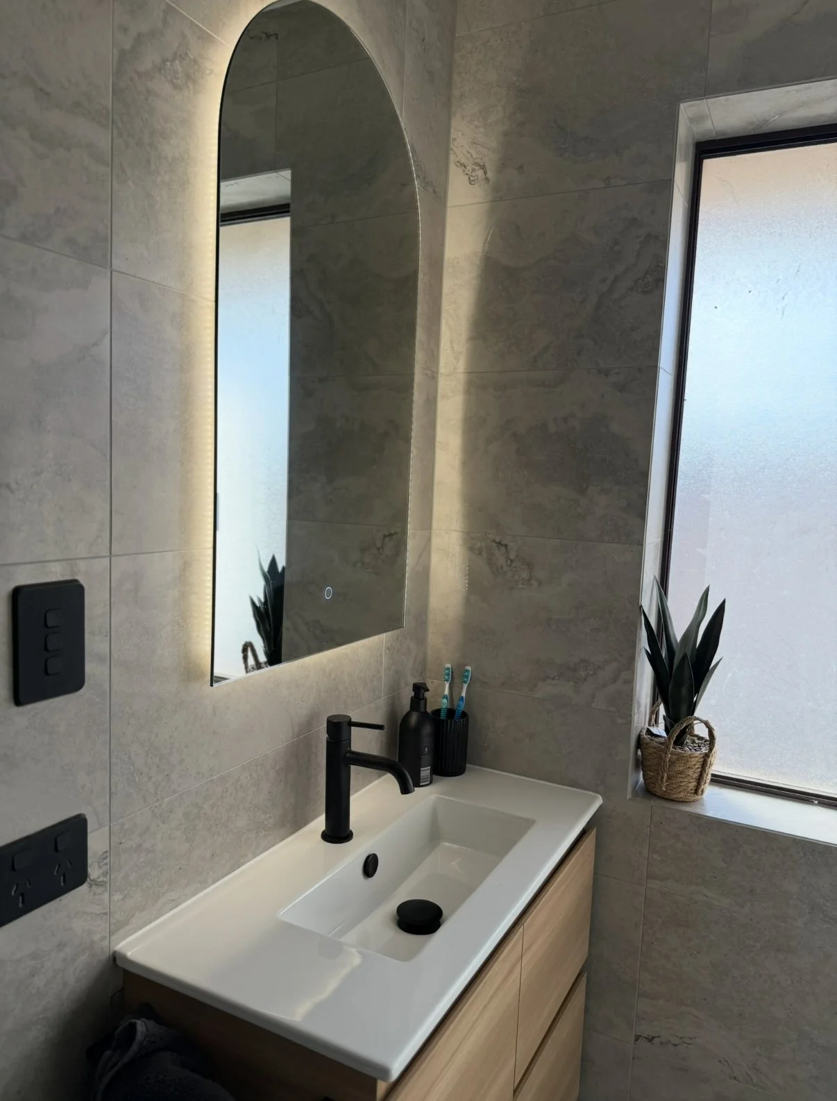 Modern bathroom with a white sink, black faucet, large mirror with LED lighting, and a window with frosted glass. A small potted plant with long green leaves is on the windowsill. Bathroom amenities such as toothbrushes and soap are on the sink.