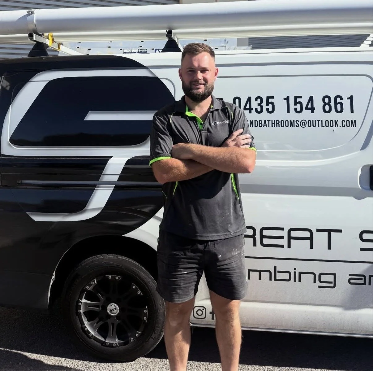 A man with a beard and short hair standing confidently with arms crossed in front of a service van. The van displays contact information and an email address. The man is dressed in a black uniform with green accents and has dirt or dust on his shorts, indicating involvement in manual or outdoor work.