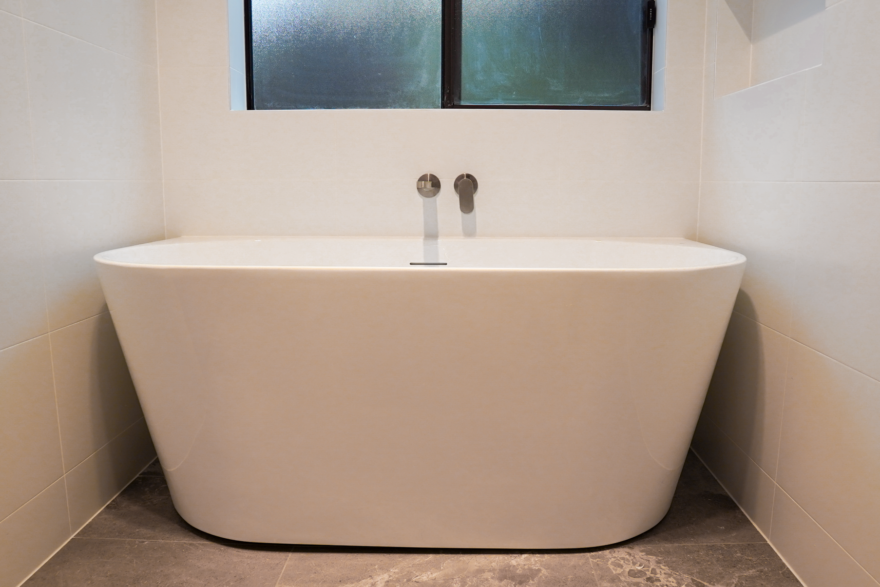 A modern white bathtub with a curved exterior, situated between beige tiled walls and a window above it, in a bathroom.