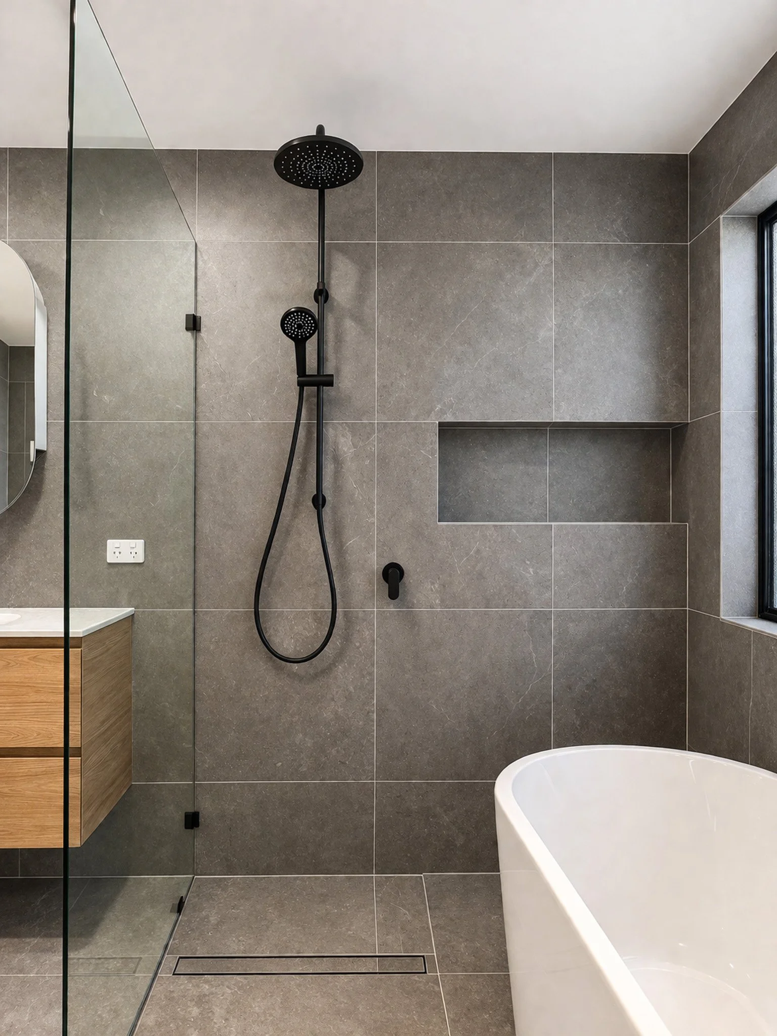 Modern shower area with a black rain shower head, a hand-held shower, gray tiled walls, a built-in niche, and a glass partition. A portion of a white bathtub is visible in the lower right corner.
