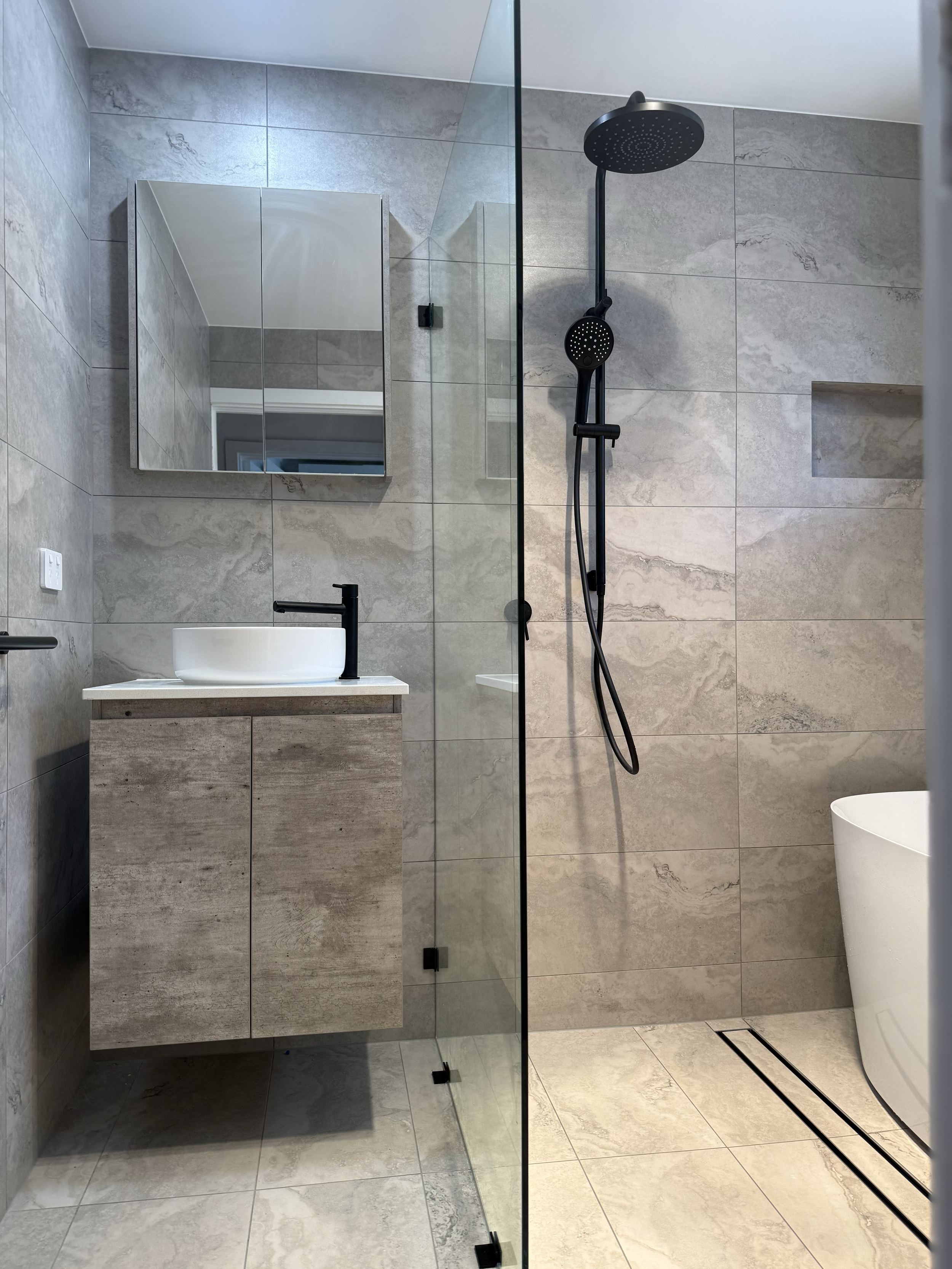 Modern bathroom with beige large tile walls and floor, glass shower partition, black rain shower head, white vessel sink with black faucet, rectangular mirror, and a white bathtub.