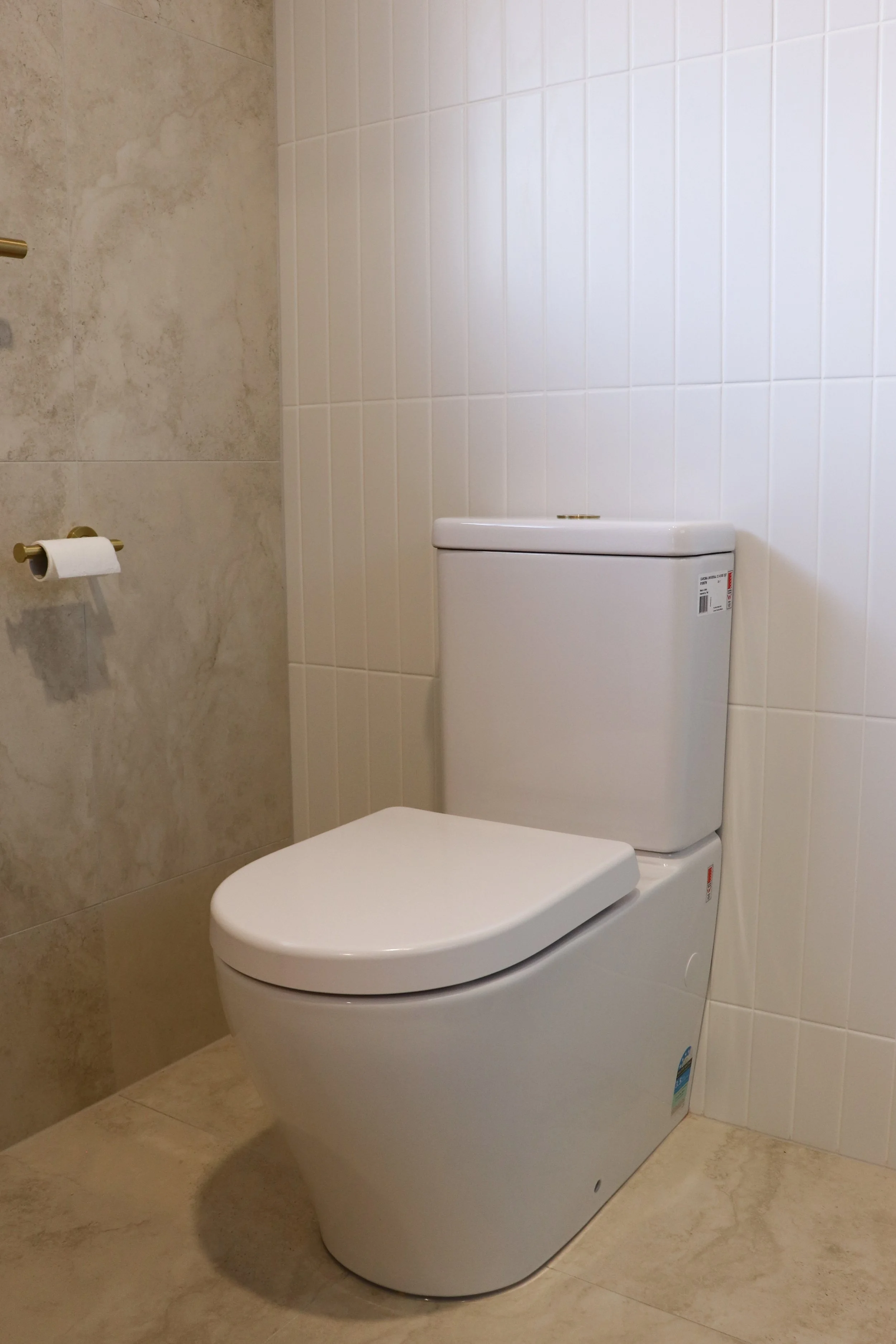 A modern indoor bathroom with a white toilet, beige tiled floor, and white tiled wall. A toilet paper holder with a roll of toilet paper is mounted on the wall to the left of the toilet.