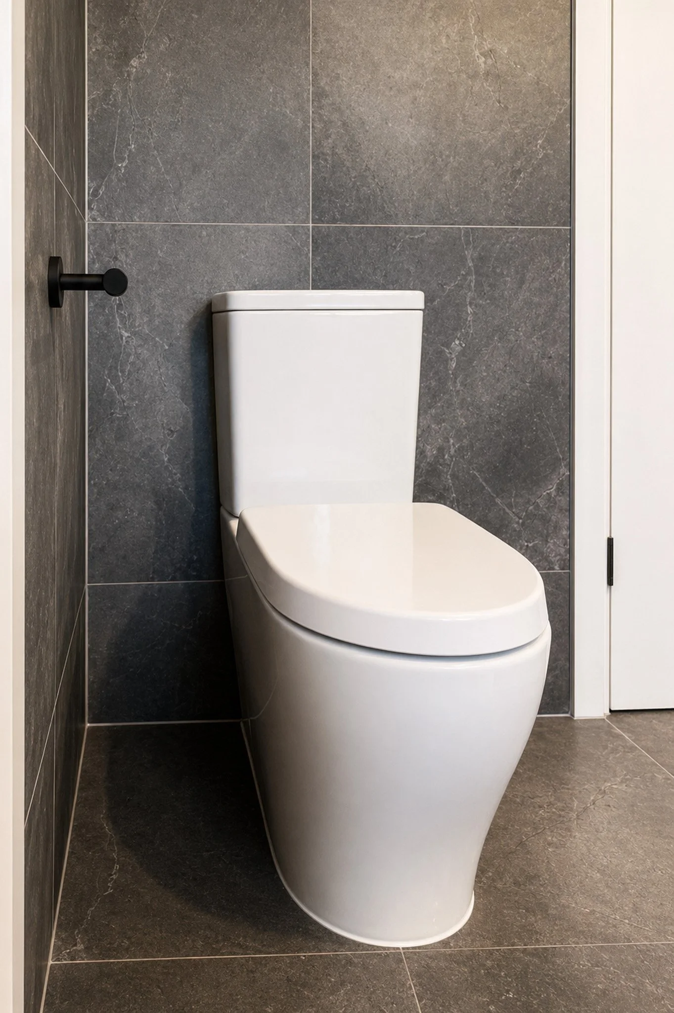Modern bathroom with a white toilet against a dark gray tiled wall and gray tiled floor. There is a black toilet paper holder on the left side.