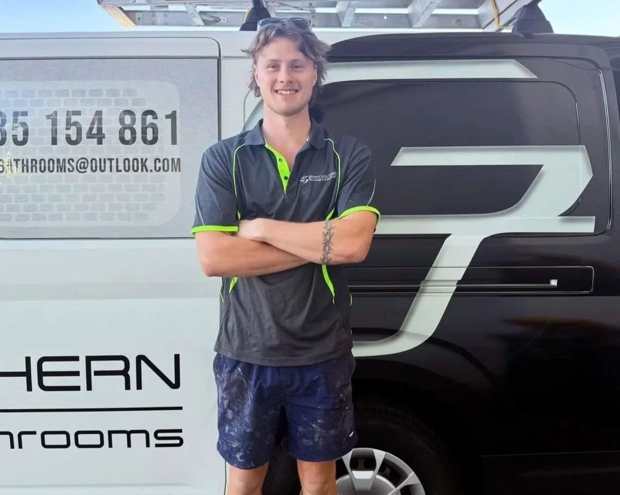 A young man with curly hair, wearing a black and green polo shirt and dark blue shorts with paint splatters, stands with arms crossed in front of a vehicle with a logo and contact information for a company called Southern Showers.