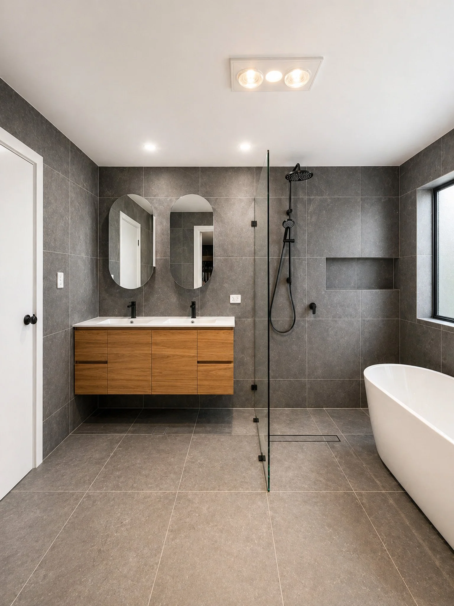 Modern bathroom with gray tiled walls and floor, double sink vanity with wood cabinet, oval mirrors, black fixtures, a separate shower area with black shower head and a separate bathtub, and a small window.
