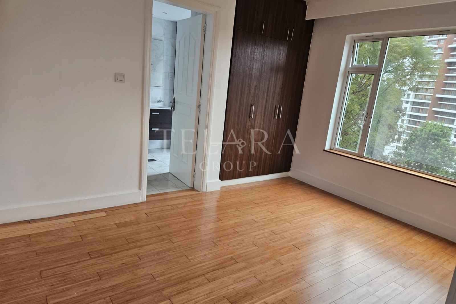 Empty room with wooden floor, large window showing trees and apartment building, built-in wooden closet, door to bathroom, white walls.