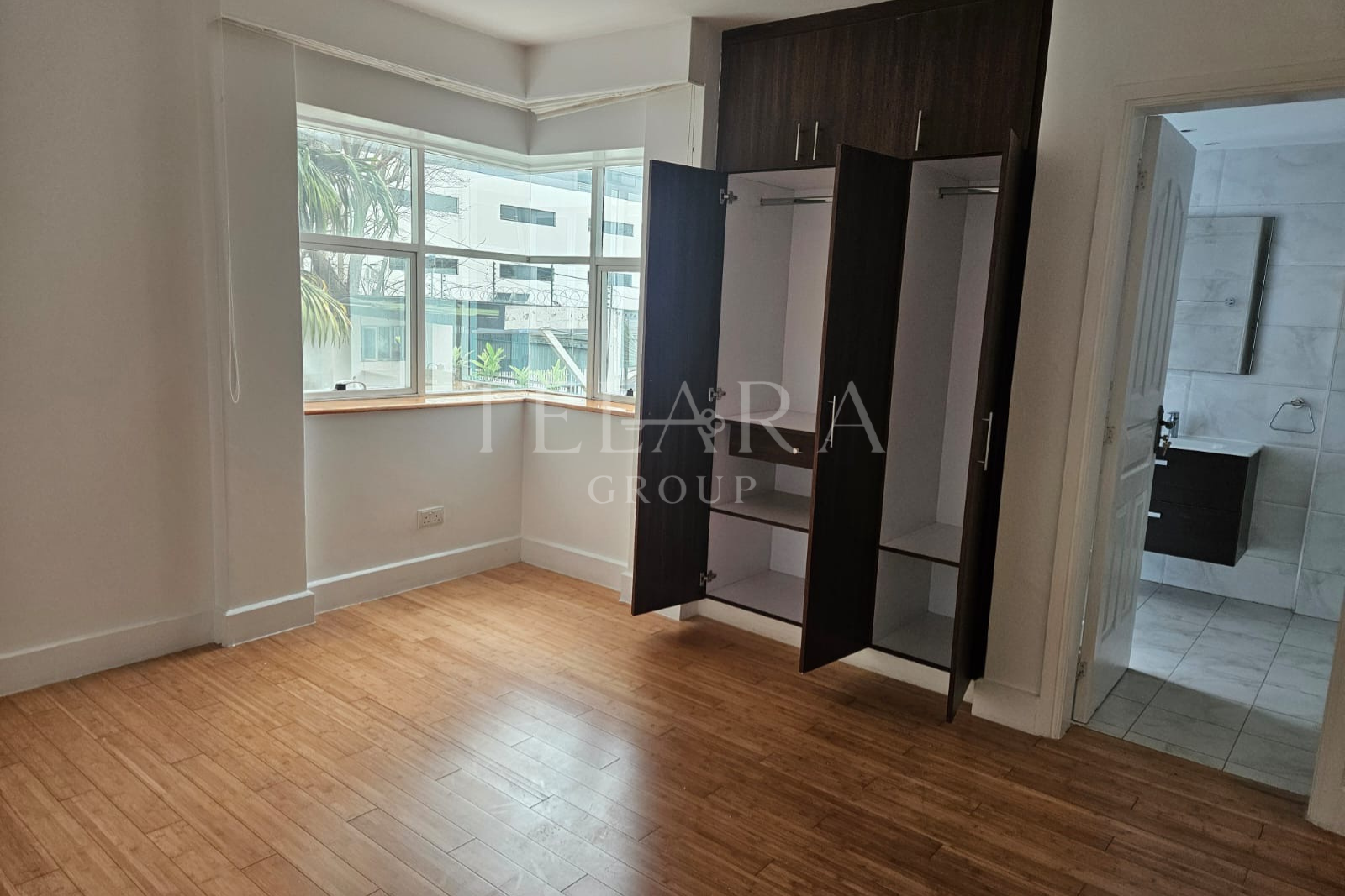 Empty room with wooden flooring, a large window with a view of neighboring buildings, and an open wardrobe with dark wood doors. A doorway to a bathroom with marble tiles and a black sink cabinet is visible.