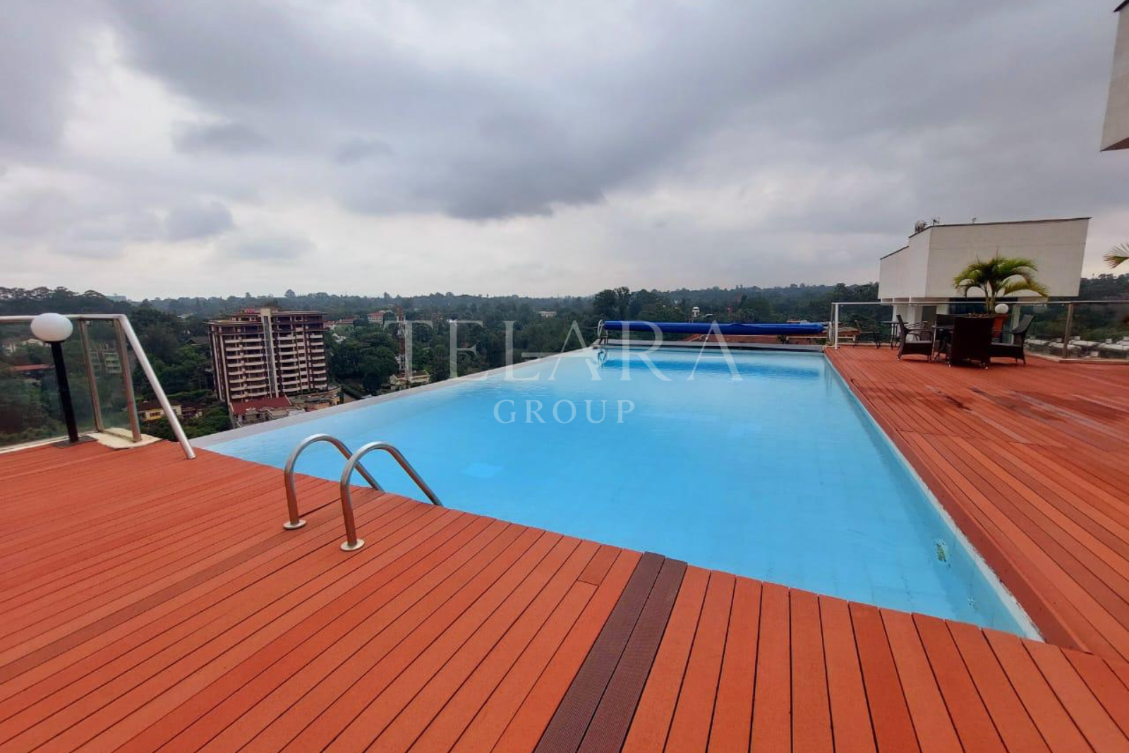 Rooftop swimming pool with a wooden deck, outdoor seating area, and city skyline in the background under cloudy sky.
