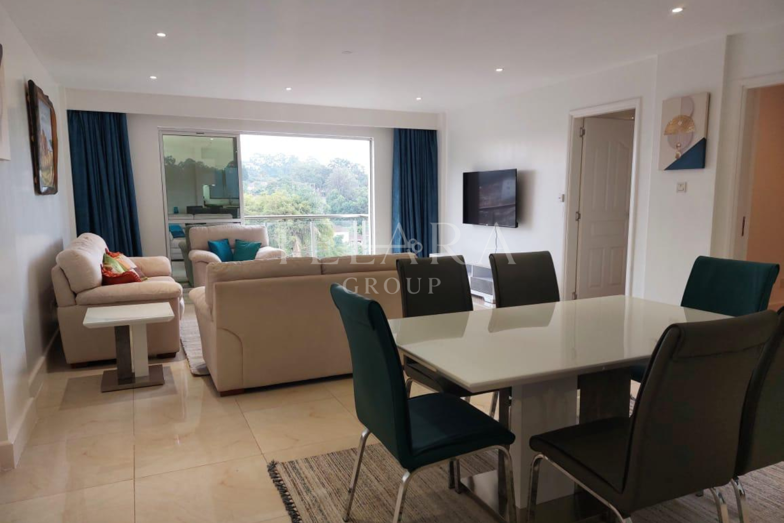 Living room with cream-colored sofas, blue pillows, a white dining table with six dark chairs, a wall-mounted TV, large window with blue curtains, and decor paintings on white walls.