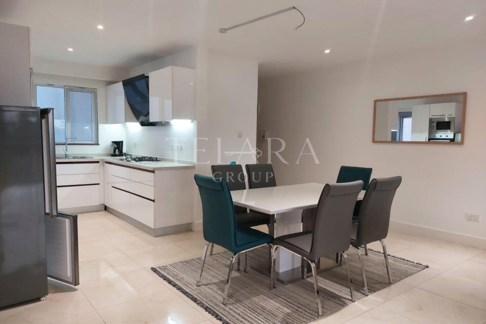 Modern open-plan kitchen and dining area with white cabinets, black range hood, gas stove, coffee maker, gray and teal chairs around a white dining table, window, and a large mirror on the wall.
