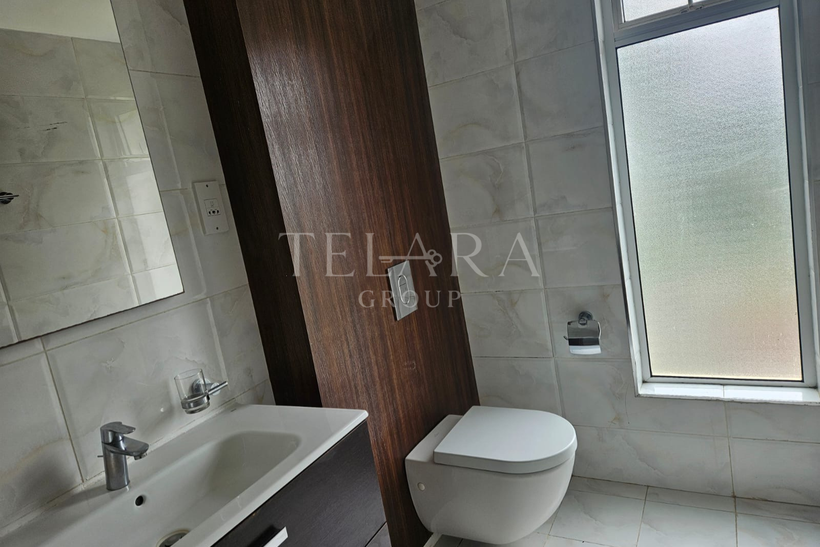 Modern bathroom with marble tile walls, a white toilet, a small white sink with a chrome faucet, a dark wood vanity, and a frosted window.