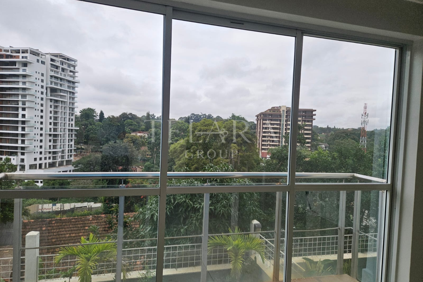 View of cityscape with tall modern buildings, trees, and a cloudy sky through a large glass window with sliding panes and a metal railing.
