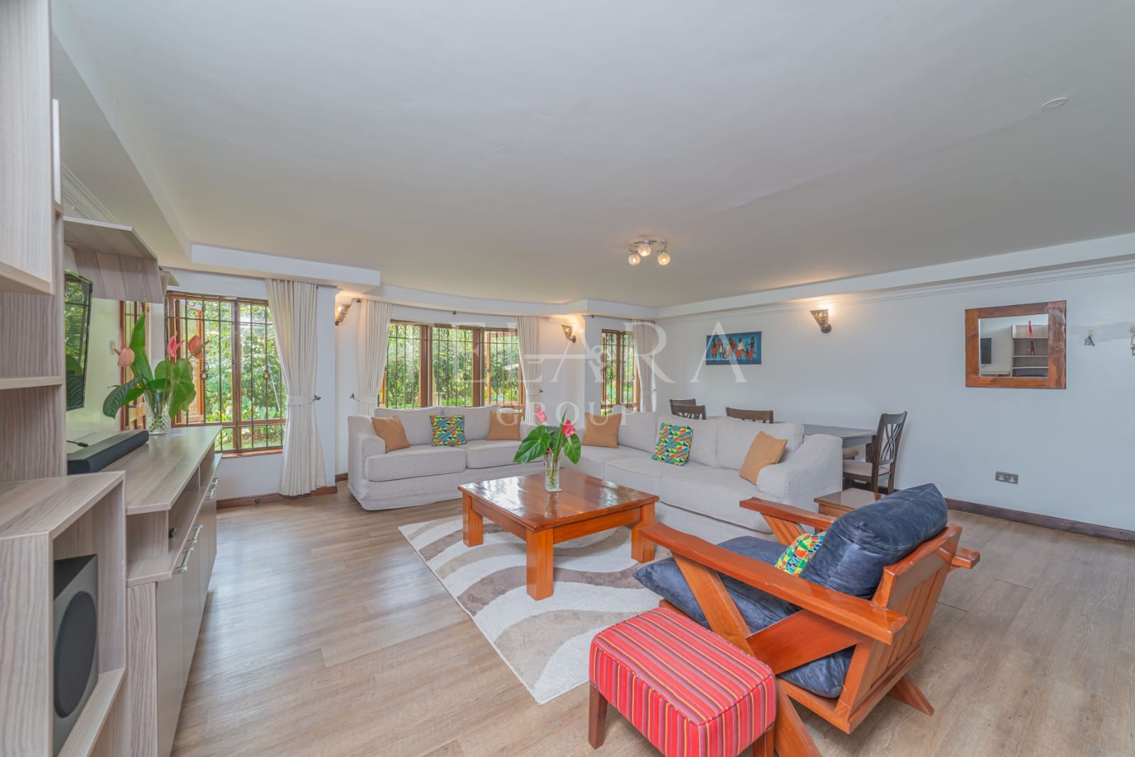Spacious living room with white sectional sofa, wooden armchair with black cushions, coffee table with a vase of pink flowers, large windows with white curtains, and hardwood flooring.