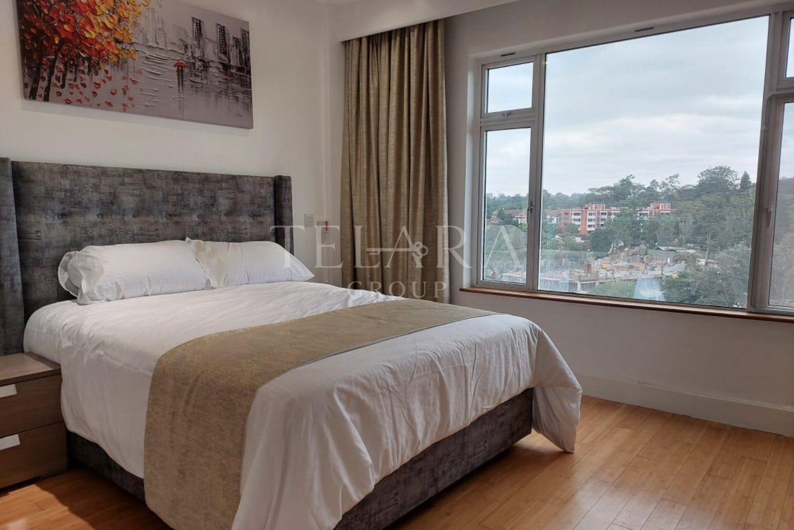 Bedroom with large window showing city view, bed with white linens and beige throw, and abstract artwork on the wall.