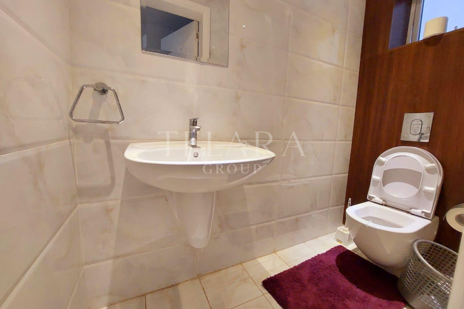 Bathroom with beige tiled walls and floor, a wall-mounted sink, a toilet with an open lid, a small window, a mirror, a purple rug, and a gray basket.
