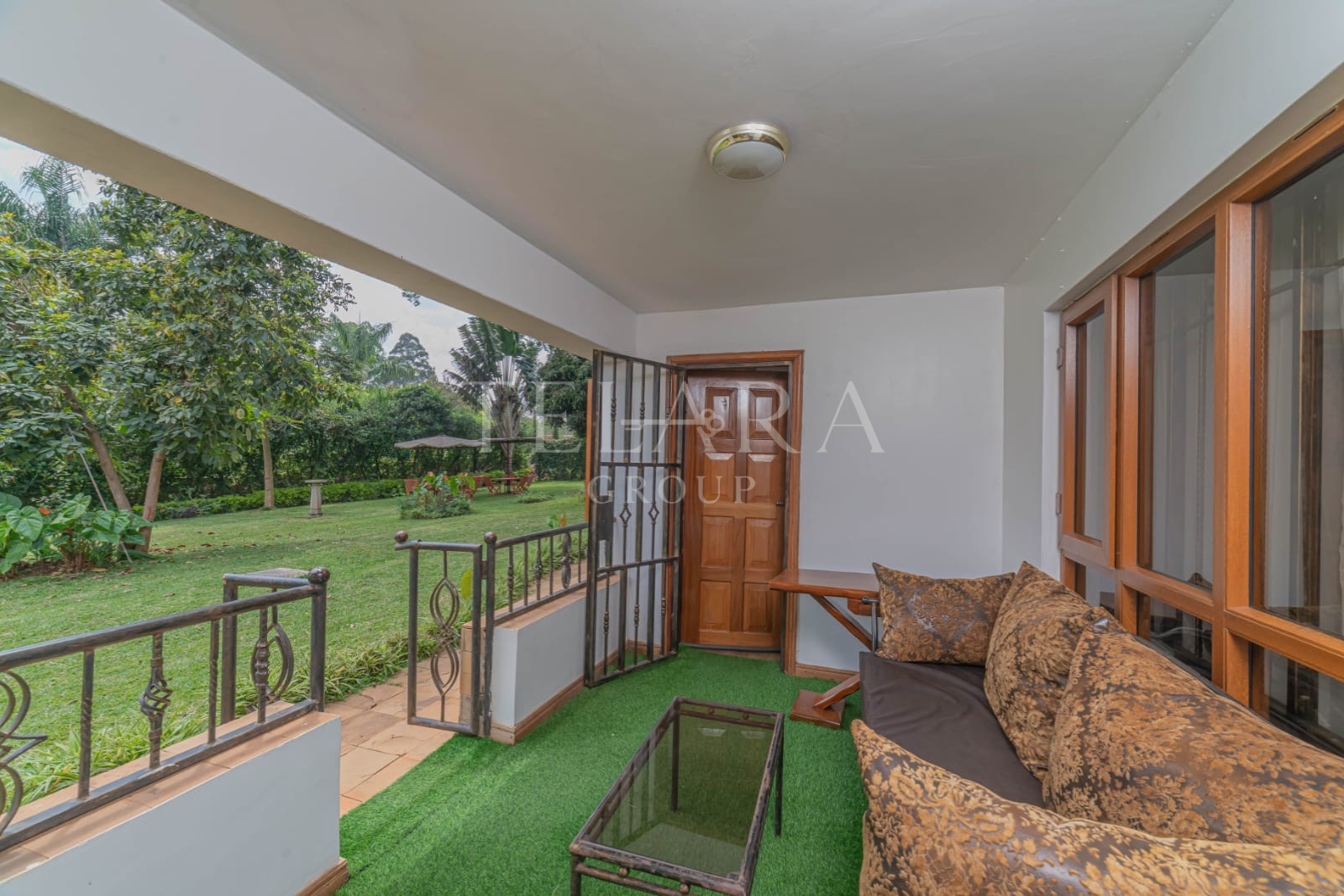 Interior view of a small porch or sunroom with a brown patterned sofa, glass-top coffee table, large windows, and a door leading outside to a green garden with trees and picnic table.