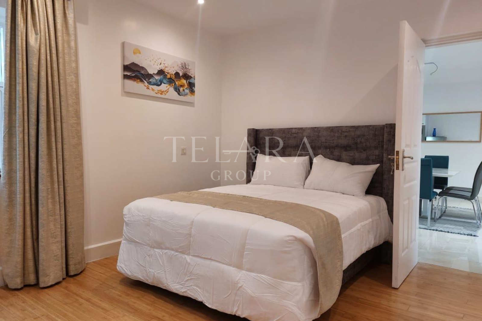 A neatly made bed with white linens, pillows, and a beige bed runner, situated in a bedroom with a painting on the wall, beige curtains, and an open door leading to a dining area.