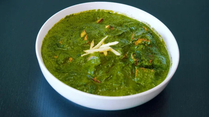 A white bowl filled with green curry garnished with sliced ginger on top, placed on a dark surface.