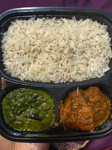 A black meal container with white rice, green curry, and a meat curry.