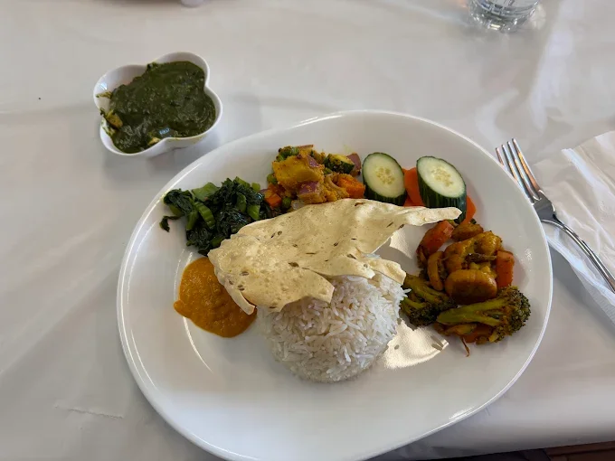 Plate of Indian curry with rice, vegetables, and a papadum, with a side of greens and a small bowl of green chutney.