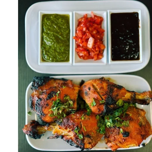 Plate of two grilled chicken drumsticks garnished with chopped herbs, served with three small bowls of green chutney, salsa, and dark sauce.