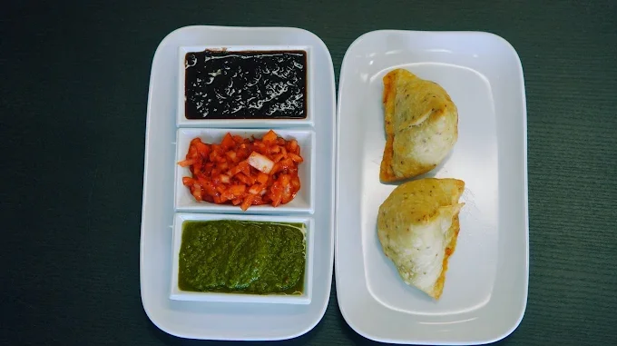 A white tray with three compartments holding dark sauce, spicy chopped vegetables, and green chutney, next to a white dish with two pieces of stuffed bread or pastry.