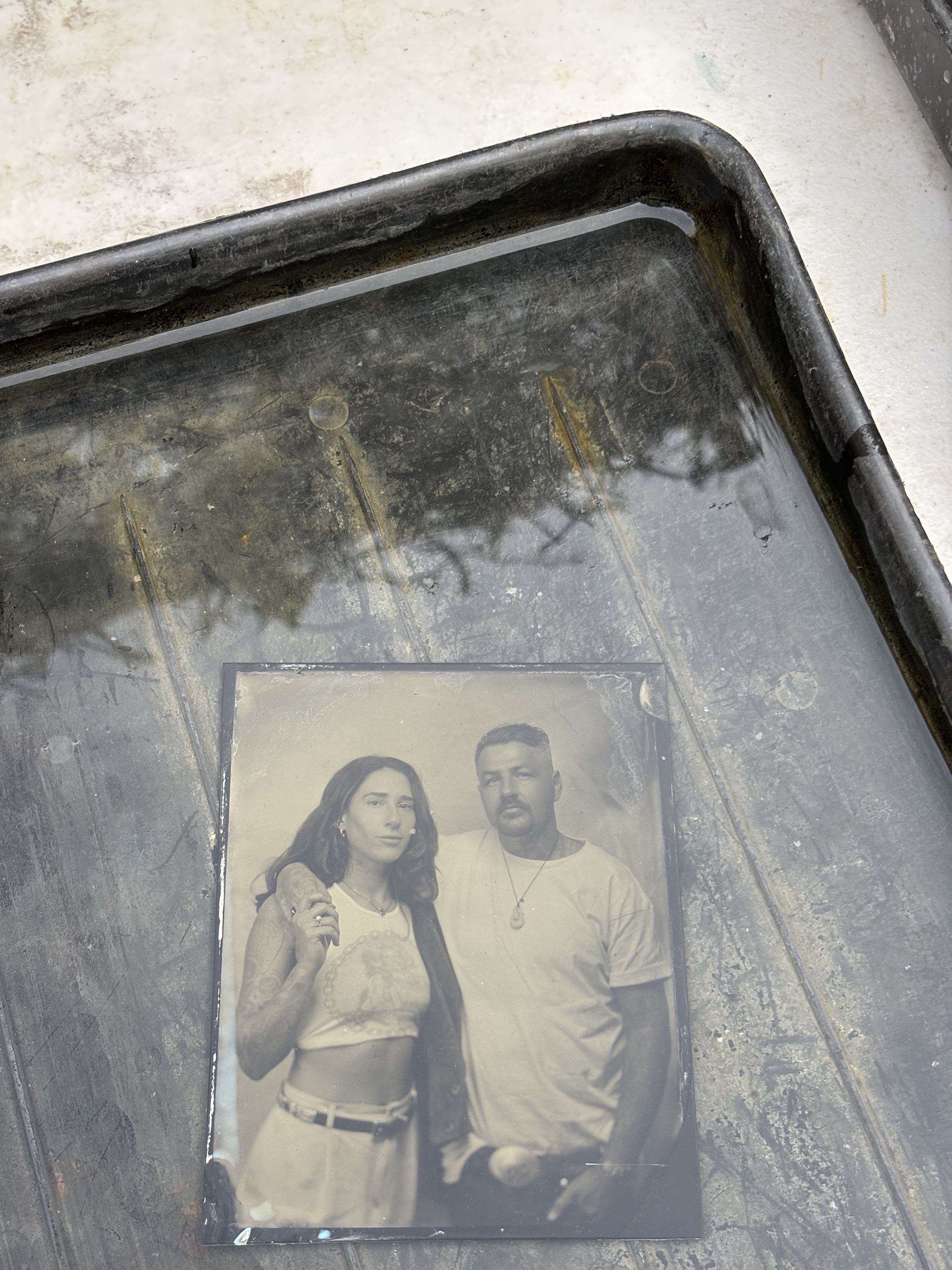 Black-and-white portrait photograph of a couple standing close together, placed in a shallow tray filled with water.