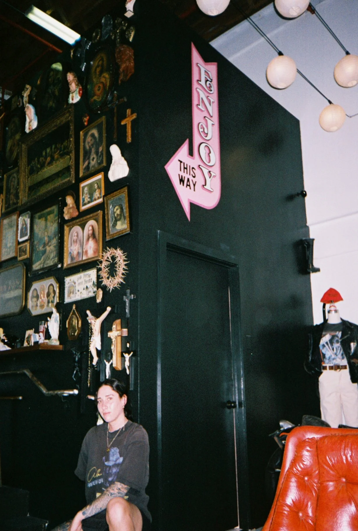 Person with visible arm tattoos sitting indoors beneath a pink “Enjoy This Way” arrow sign, surrounded by framed religious artwork and vintage decor.