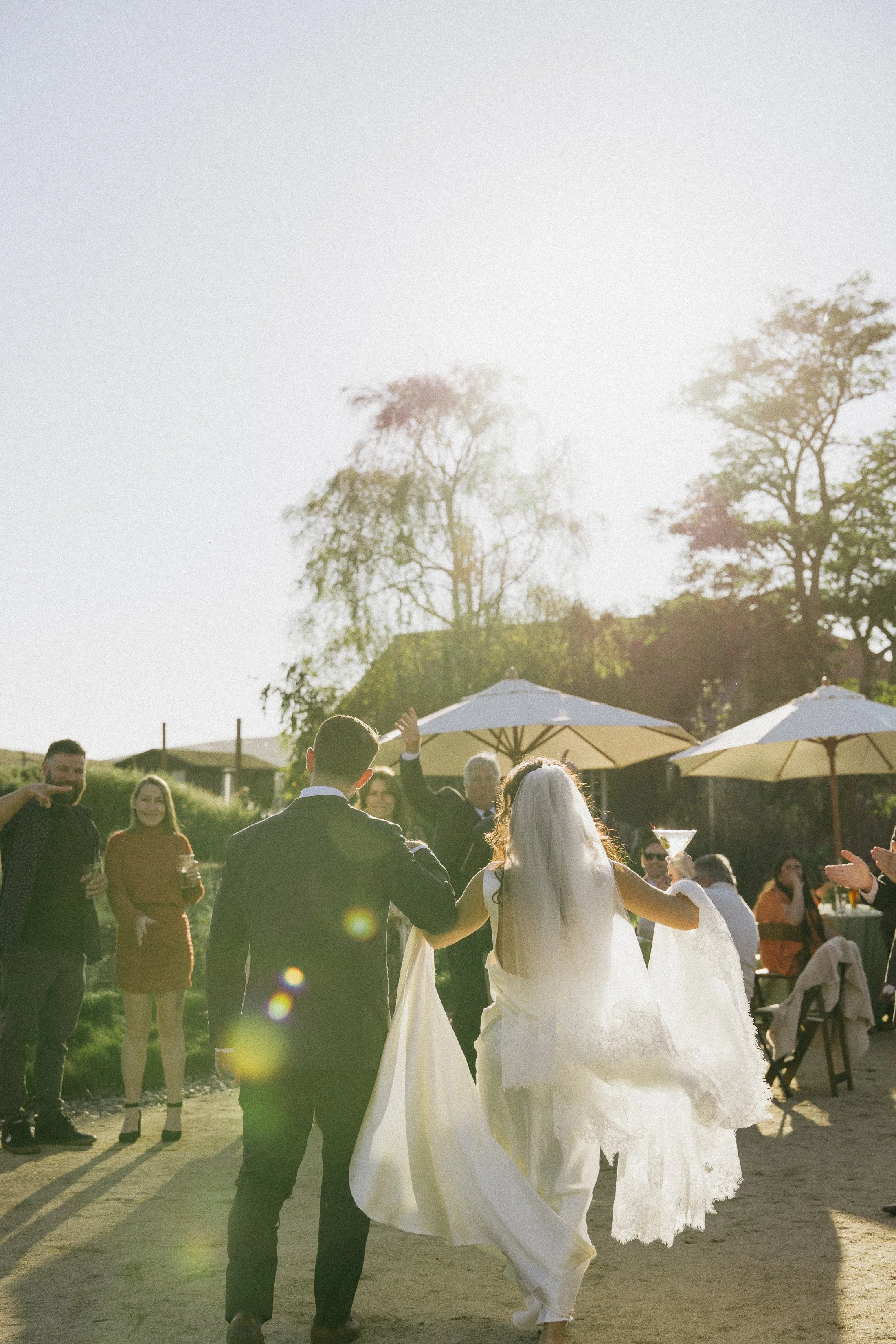 A bride and groom walk hand in hand outdoors during their wedding reception, backlit by warm golden sunlight. Guests stand on either side clapping and smiling, with white patio umbrellas and trees in the background.