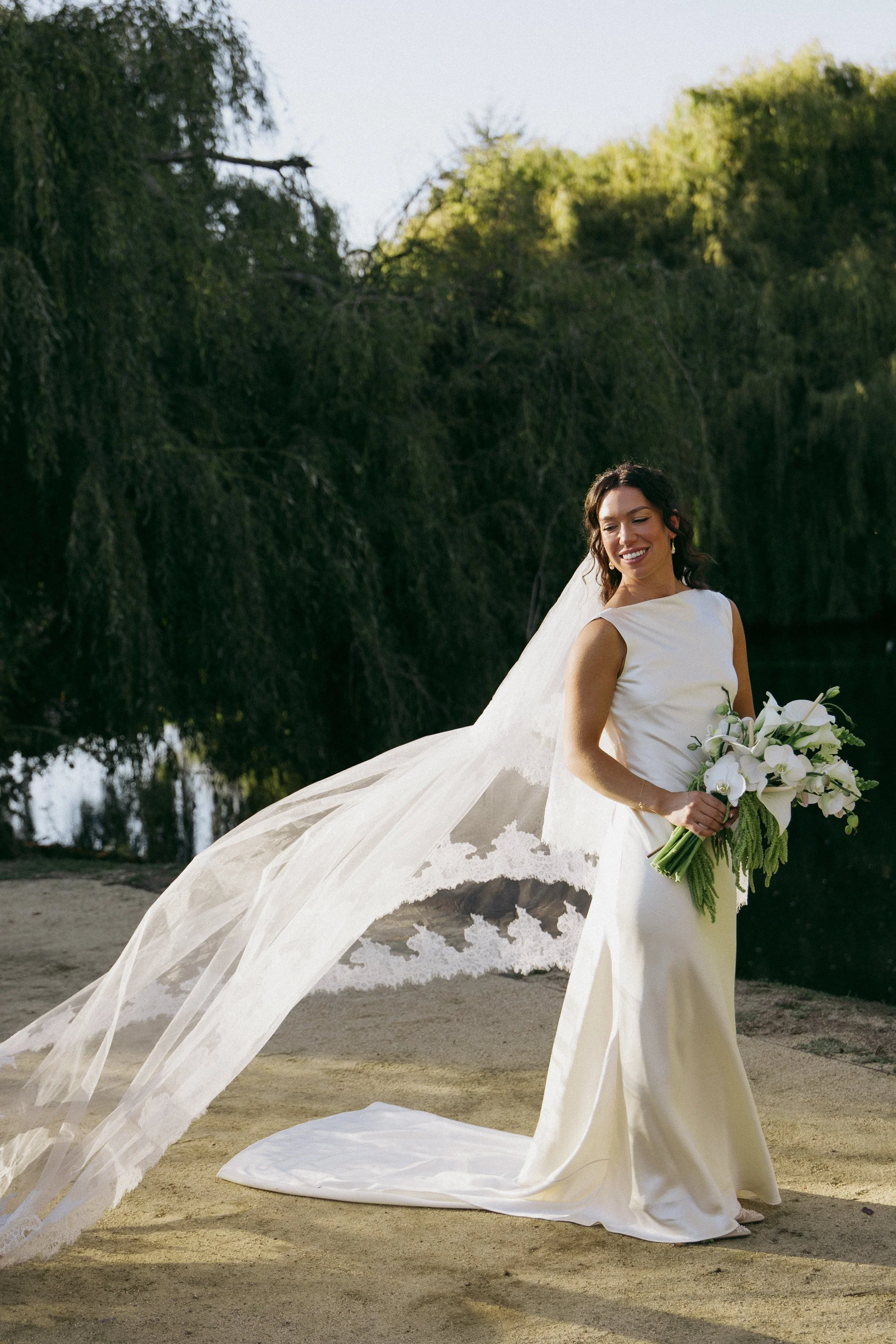 Bride Outdoors by the Water
A bride stands gracefully outdoors near a calm body of water, with lush green trees draping in the background. She wears a sleek white gown and holds a bouquet of white flowers with cascading greenery. Her long veil flows 