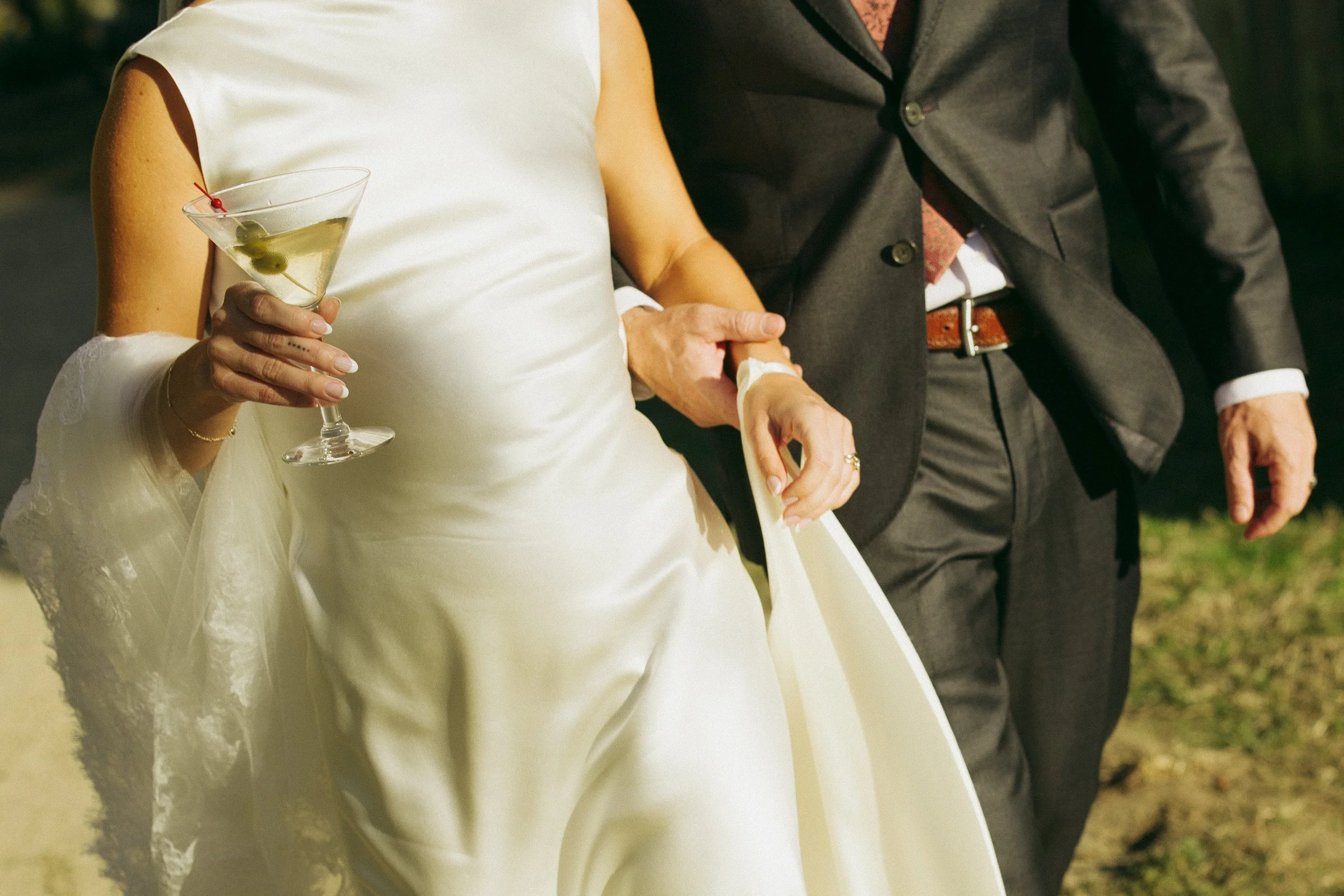 A close-up view of the bride and groom walking hand in hand outdoors. The bride holds a martini glass garnished with olives and wears a fitted white dress with a lace veil draped over her arm. The groom wears a dark suit with a tie, and warm evening 