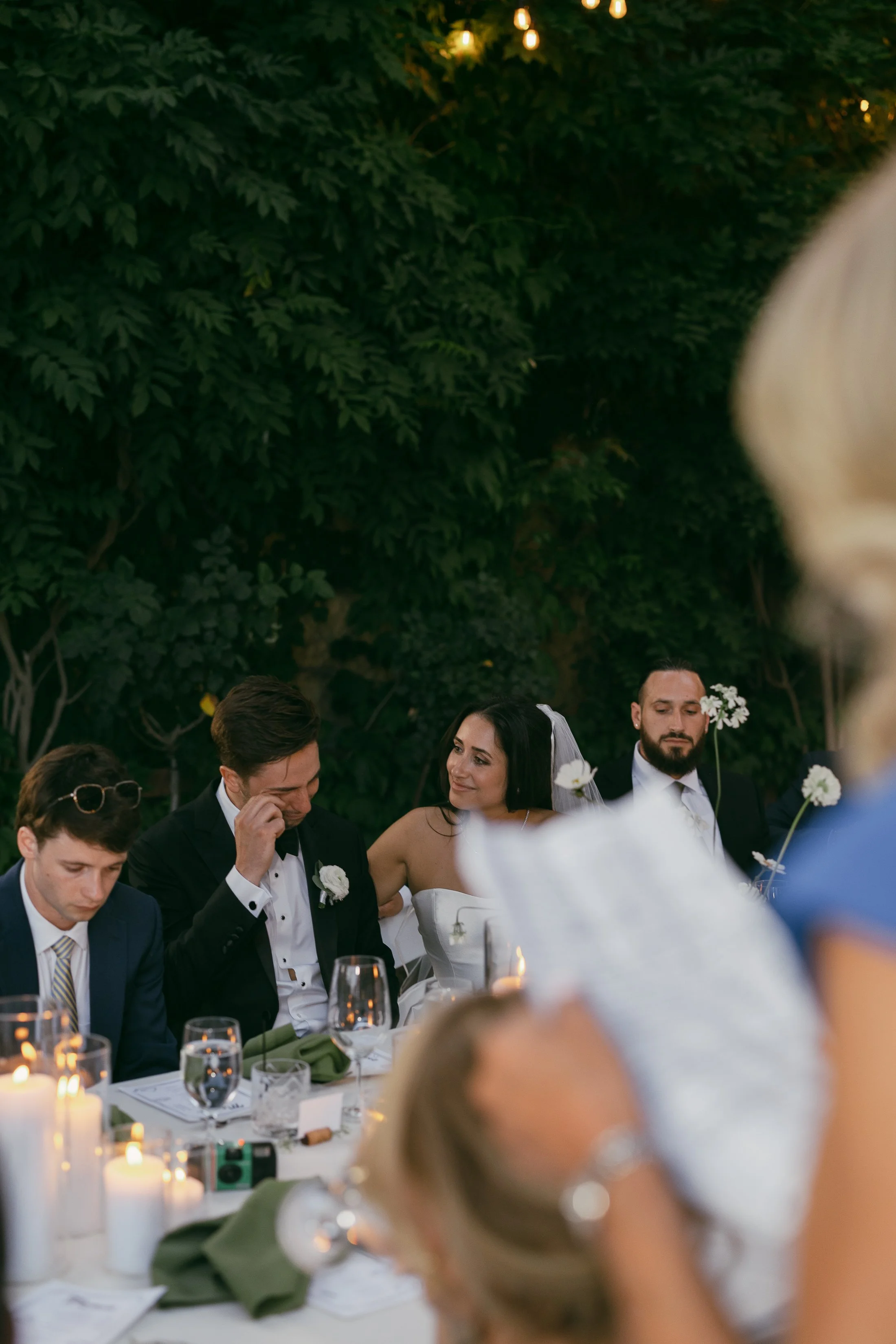 At an outdoor wedding reception, the bride sits beside the groom at a candlelit table surrounded by greenery. She smiles warmly at him as he wipes away a tear, appearing emotional during a speech. Guests in formal attire sit nearby, and soft string l