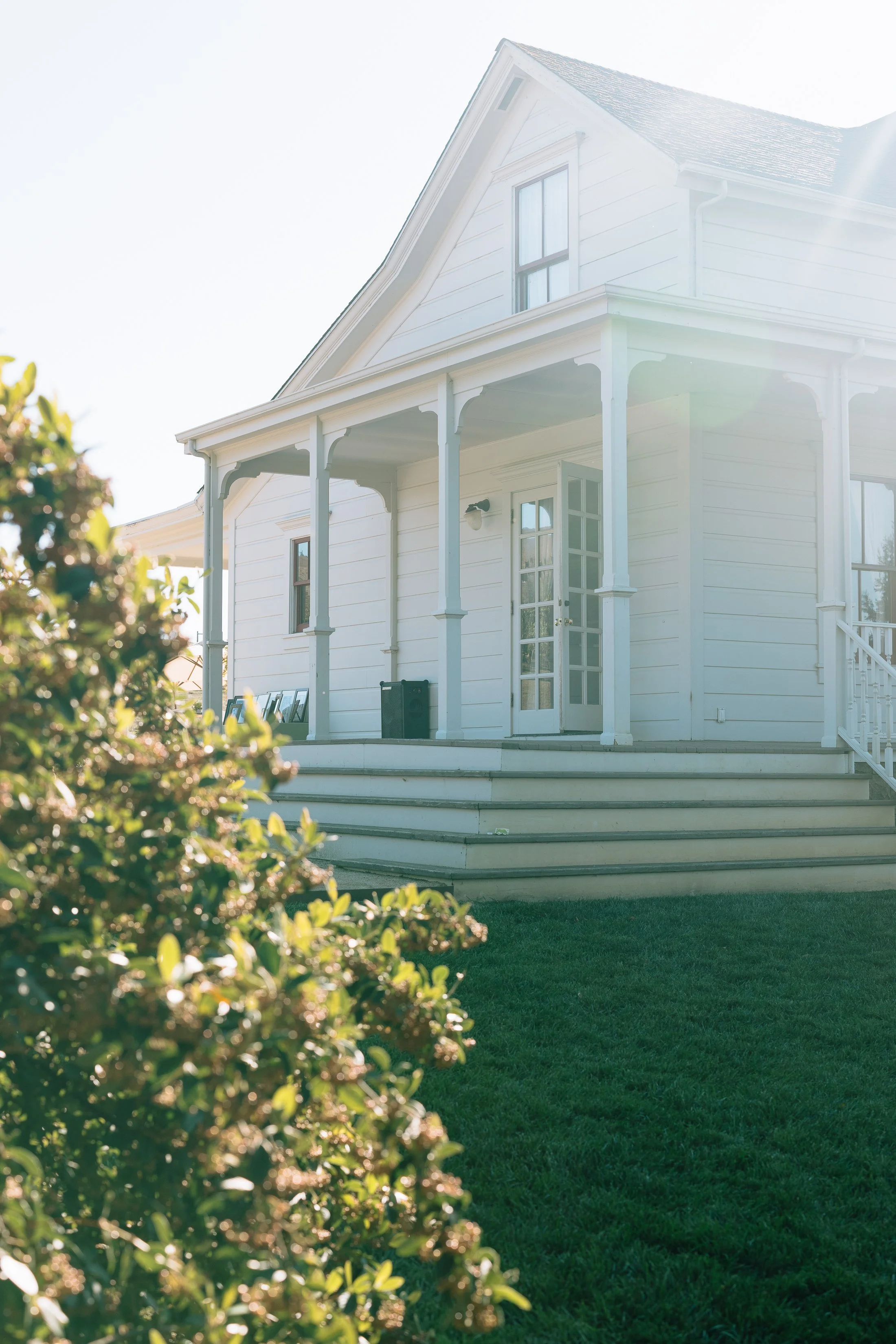 White Cottage Exterior
A charming white cottage-style house sits in soft, glowing sunlight. The home features a covered front porch with delicate trim details and pale blue-gray columns. Wide steps lead up to French doors, and neatly trimmed greenery