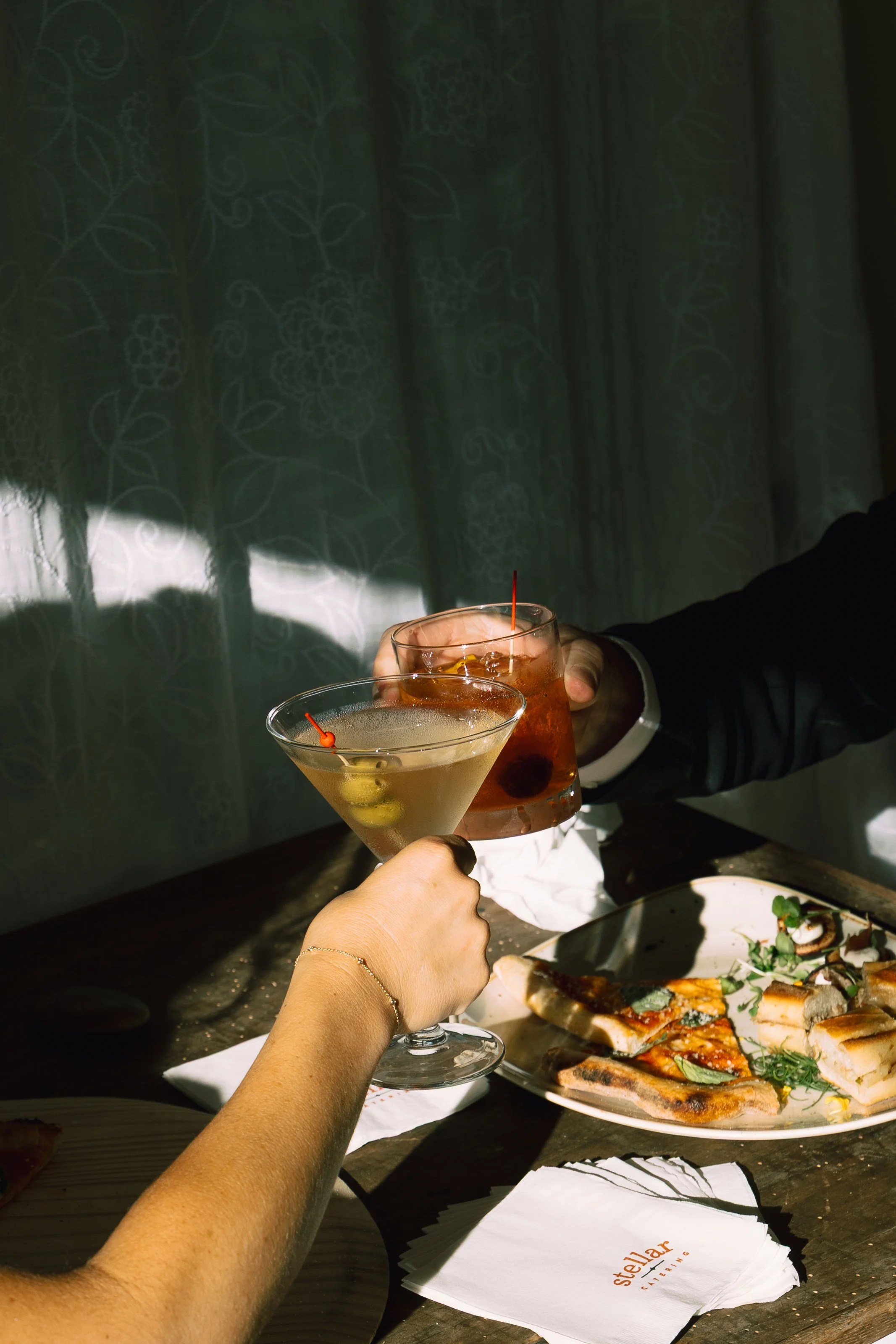 2. Cocktails and Appetizers Toast
Two people clink cocktails over a rustic wooden table bathed in warm, directional light. One hand holds a martini with olives, while the other holds an amber-colored drink with a cherry garnish. On the table are smal