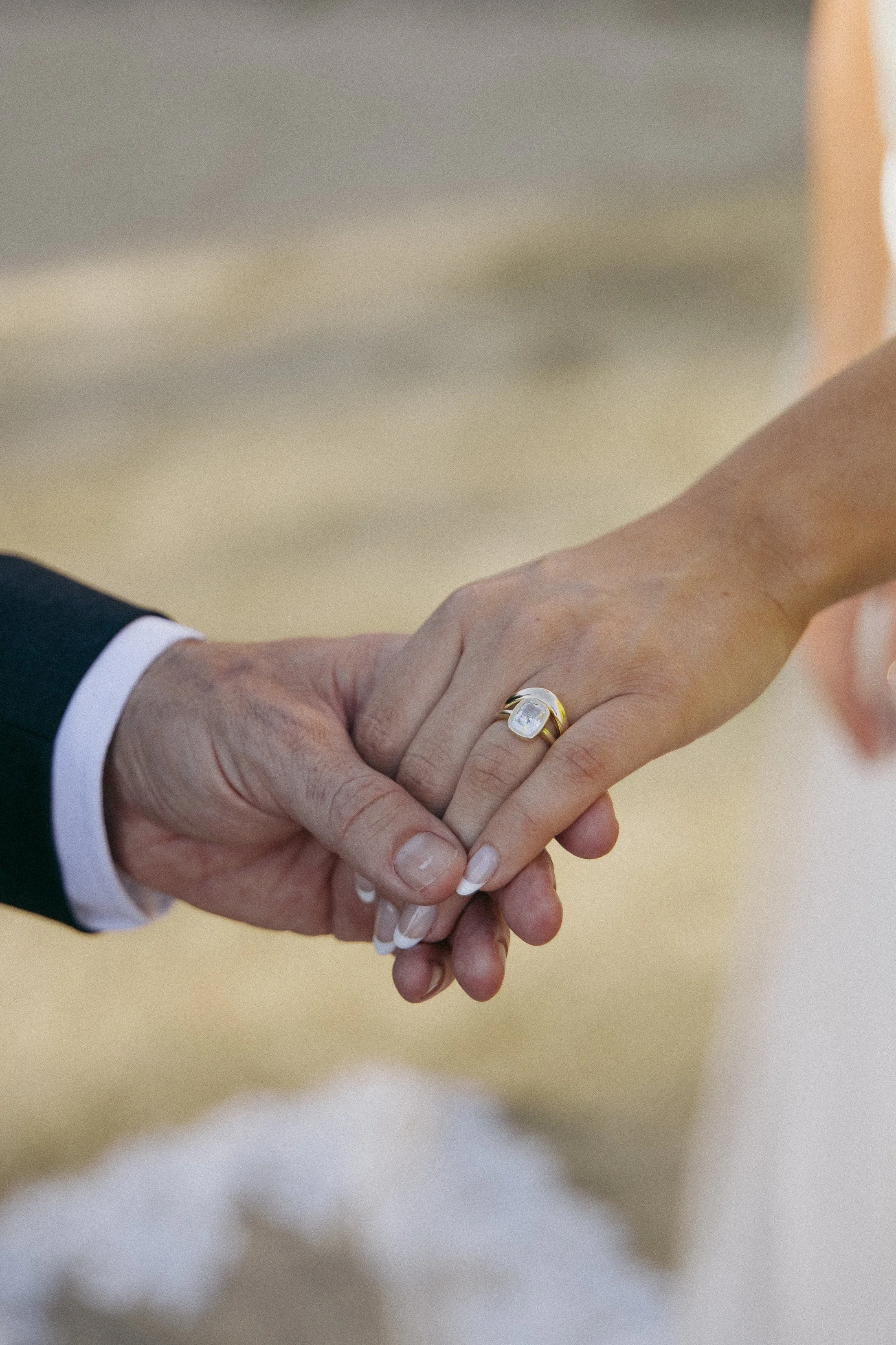 Close-up of a bride and groom holding hands, highlighting the bride’s gold ring with a large oval gemstone and her French manicure.