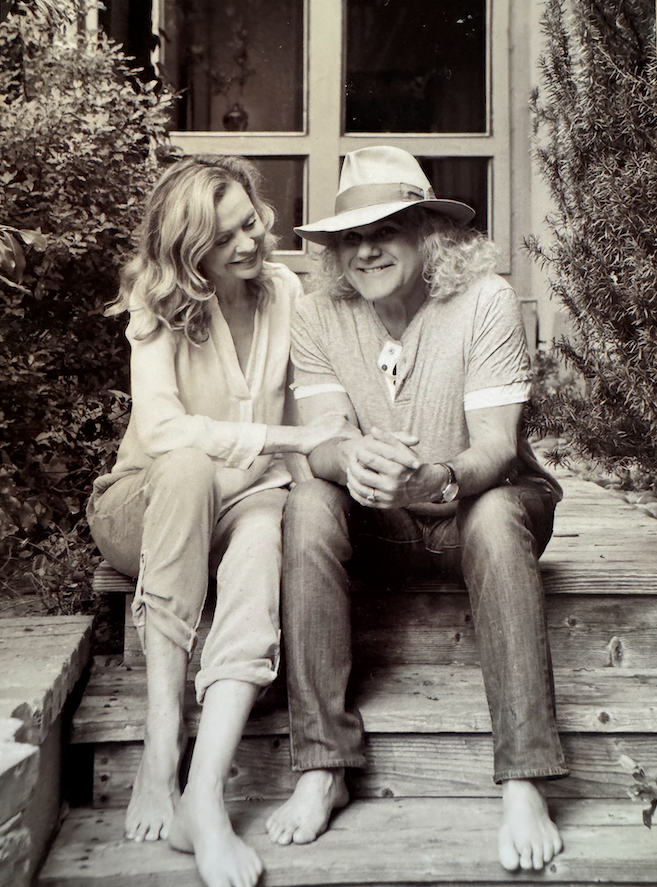 A woman and a man sitting on wooden steps outdoors, both barefoot, smiling, with the woman holding the man's arm. The woman has wavy blonde hair and is wearing a light-colored blouse and pants. The man is wearing a hat, casual t-shirt, and jeans, with long curly hair and glasses.