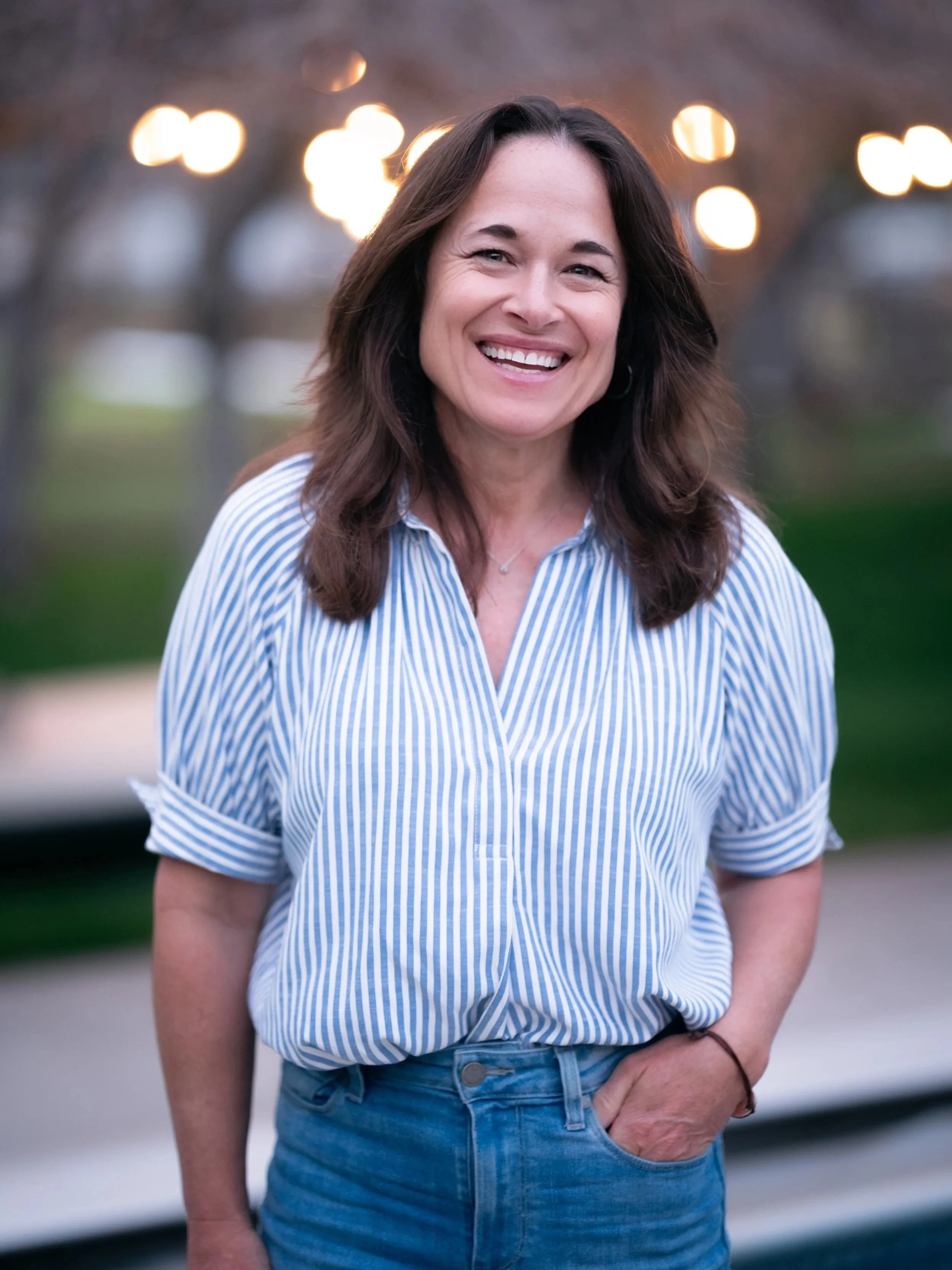 A woman smiling outdoors at dusk, wearing a blue and white striped shirt and blue jeans, with blurred lights and trees in the background.