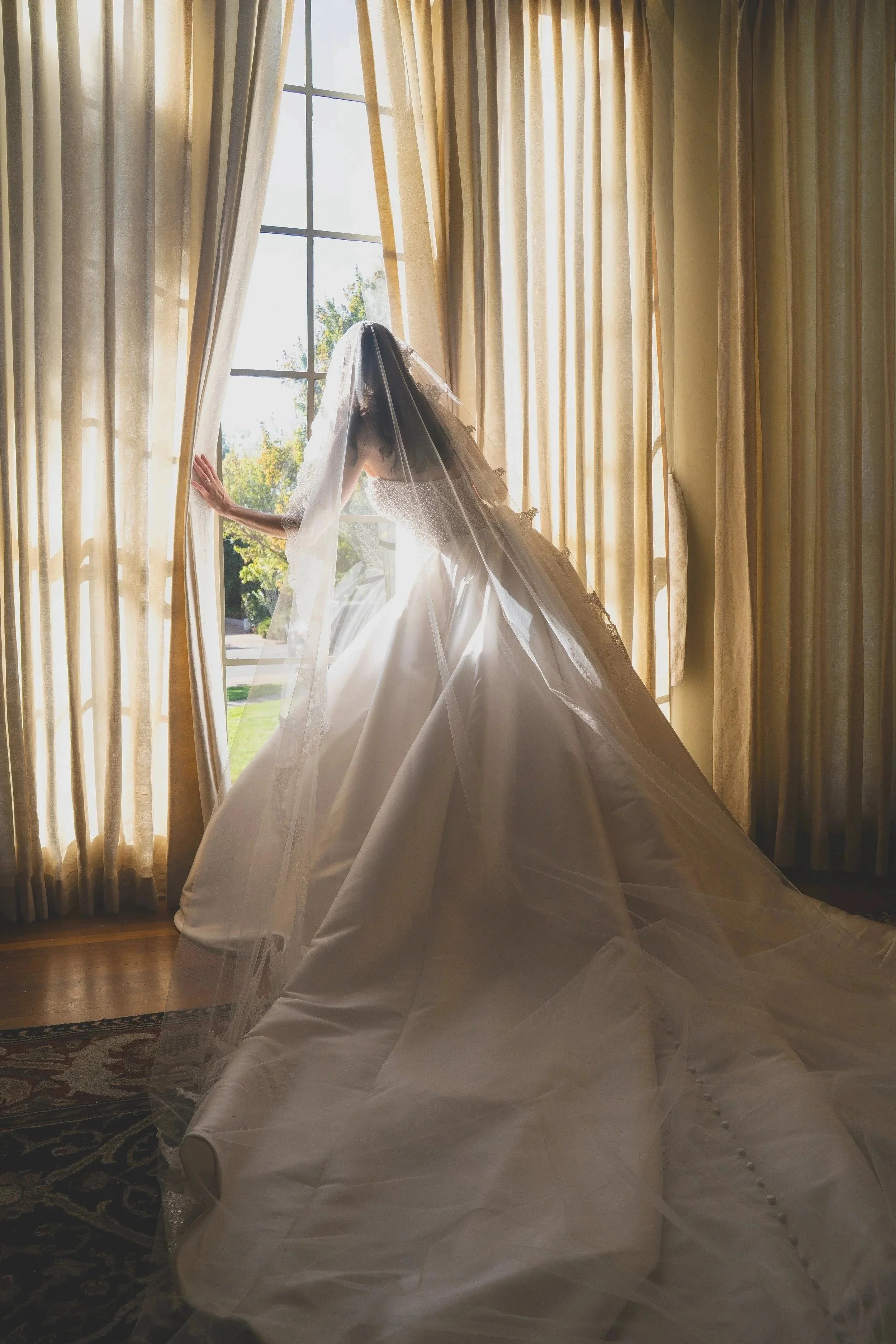 A bride in a wedding dress standing by an open window with curtains, looking outside and holding the curtain with one hand, sunlight streaming into the room.