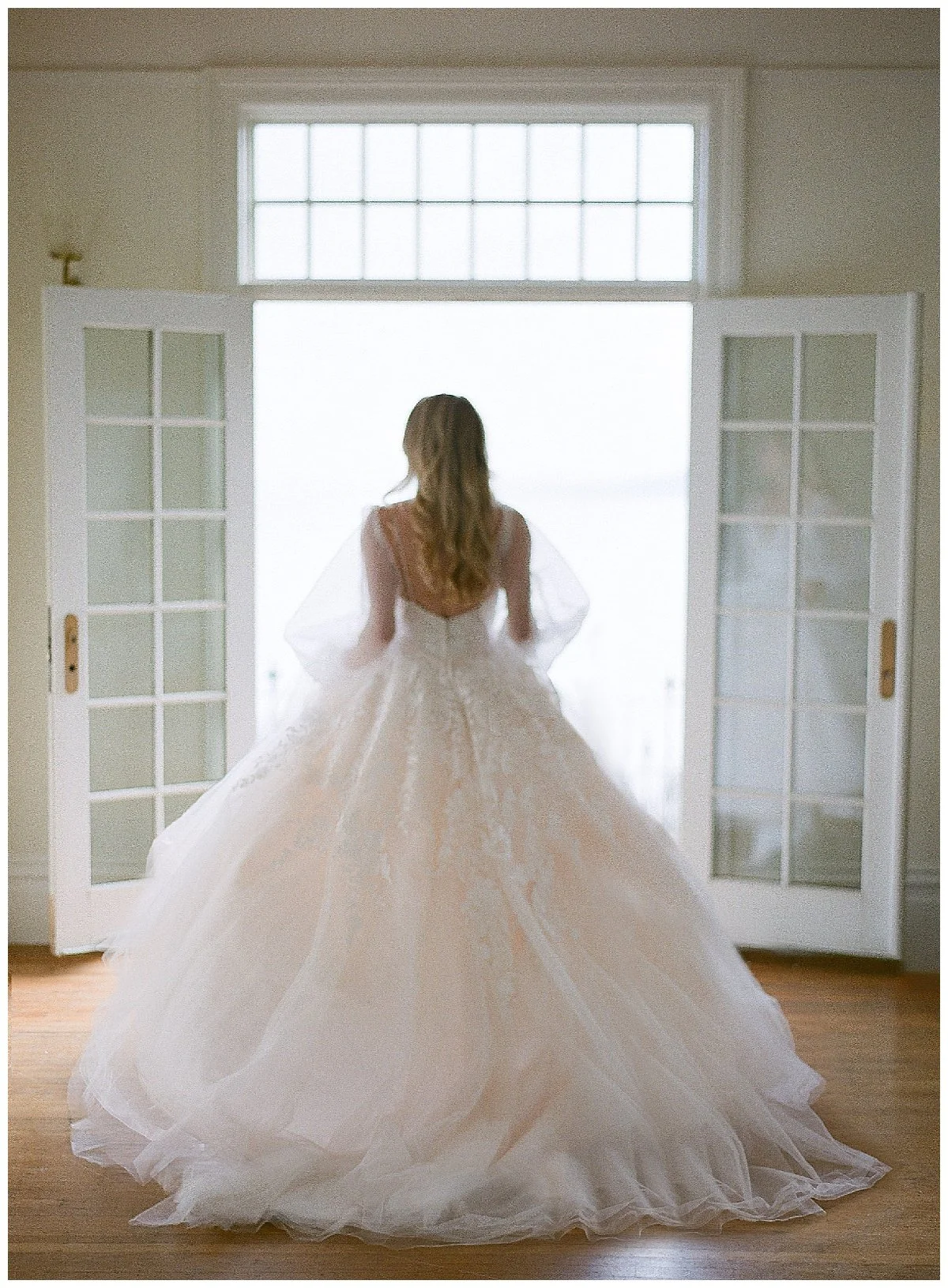 A bride in a wedding dress standing in front of open glass doors with outside bright light shining through.