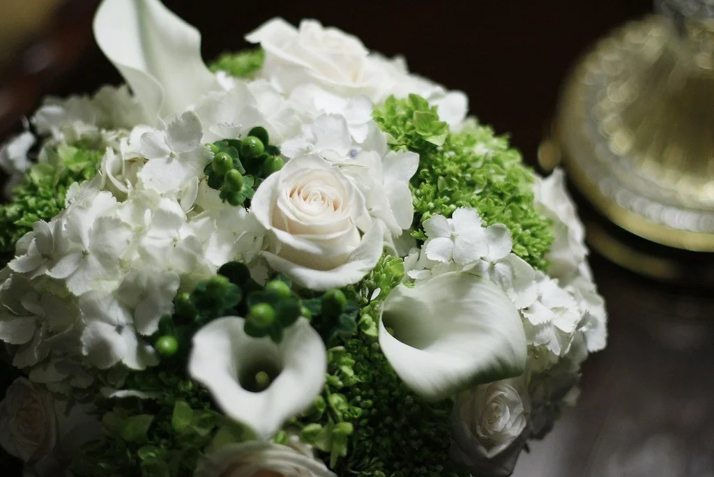 A bouquet of white flowers, including roses, hydrangeas, and calla lilies, next to a gold-colored decorative object.