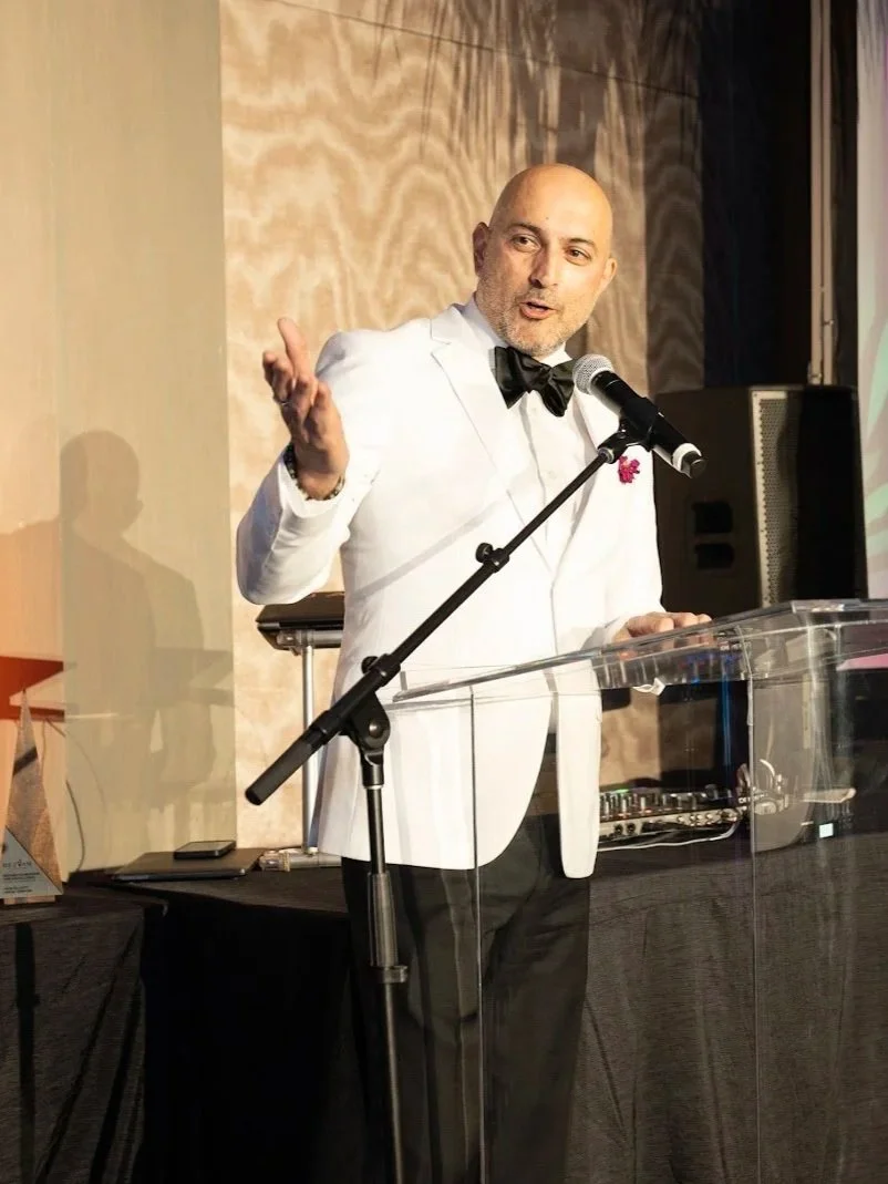 A man in a white tuxedo jacket with a black bow tie is speaking into a microphone at a podium during a formal event.