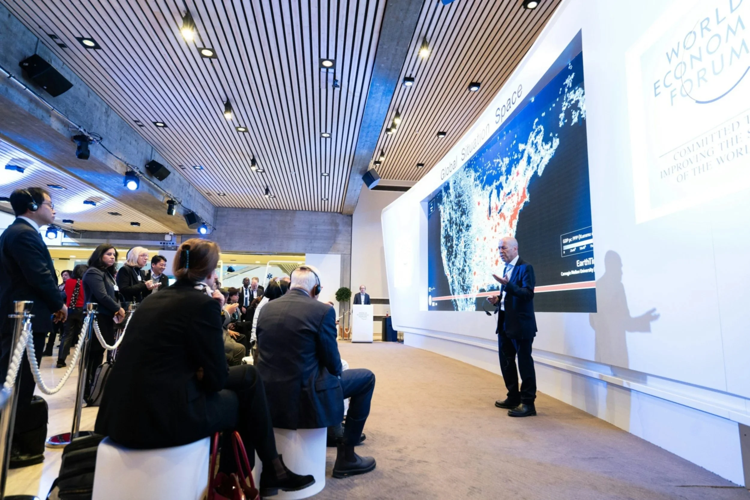 A man giving a presentation at a conference, with a large screen displaying a map of the United States titled 'Global Situation Space.' Audience members are seated and listening, some wearing headsets, in a modern conference room.