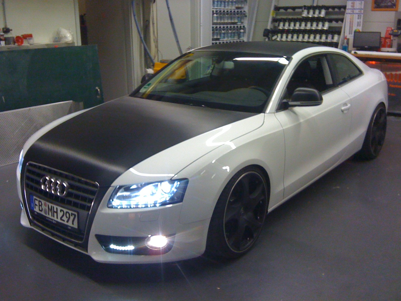 A white Audi car with a black hood and black wheels parked inside a garage or workshop with tools and equipment in the background.
