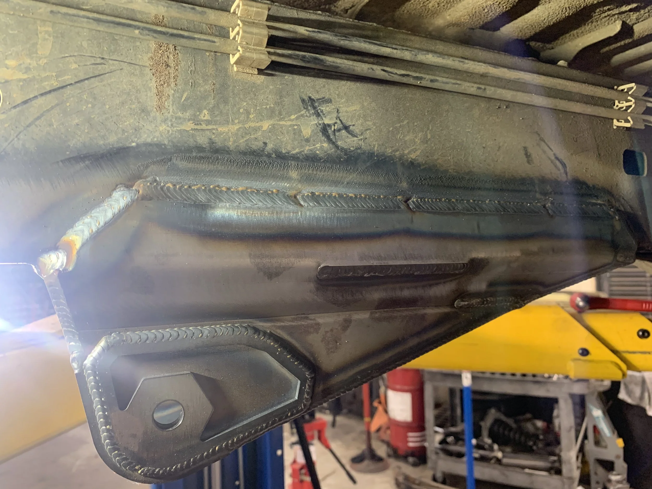 Close-up of a welded metal component, with visible weld beads and a bolt hole, in a workshop setting.