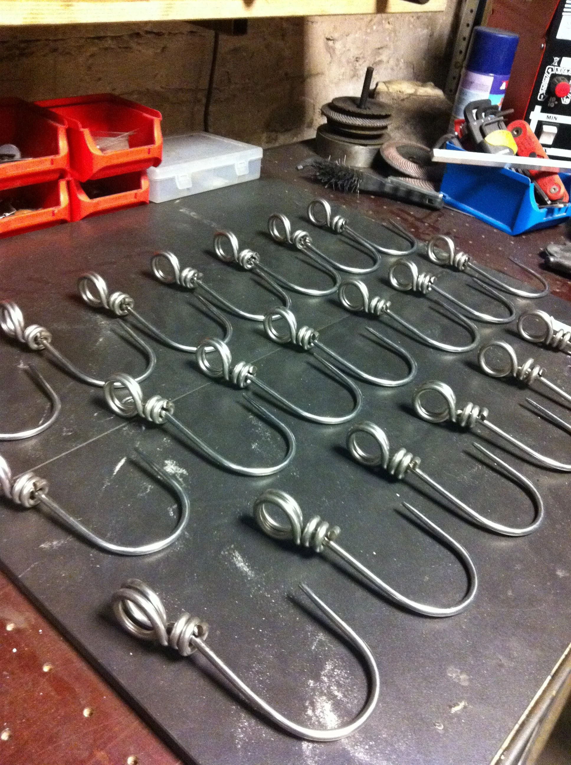 A workshop workbench with multiple metal U-bolt fasteners laid out in rows, surrounded by various tools and supplies for metalworking.