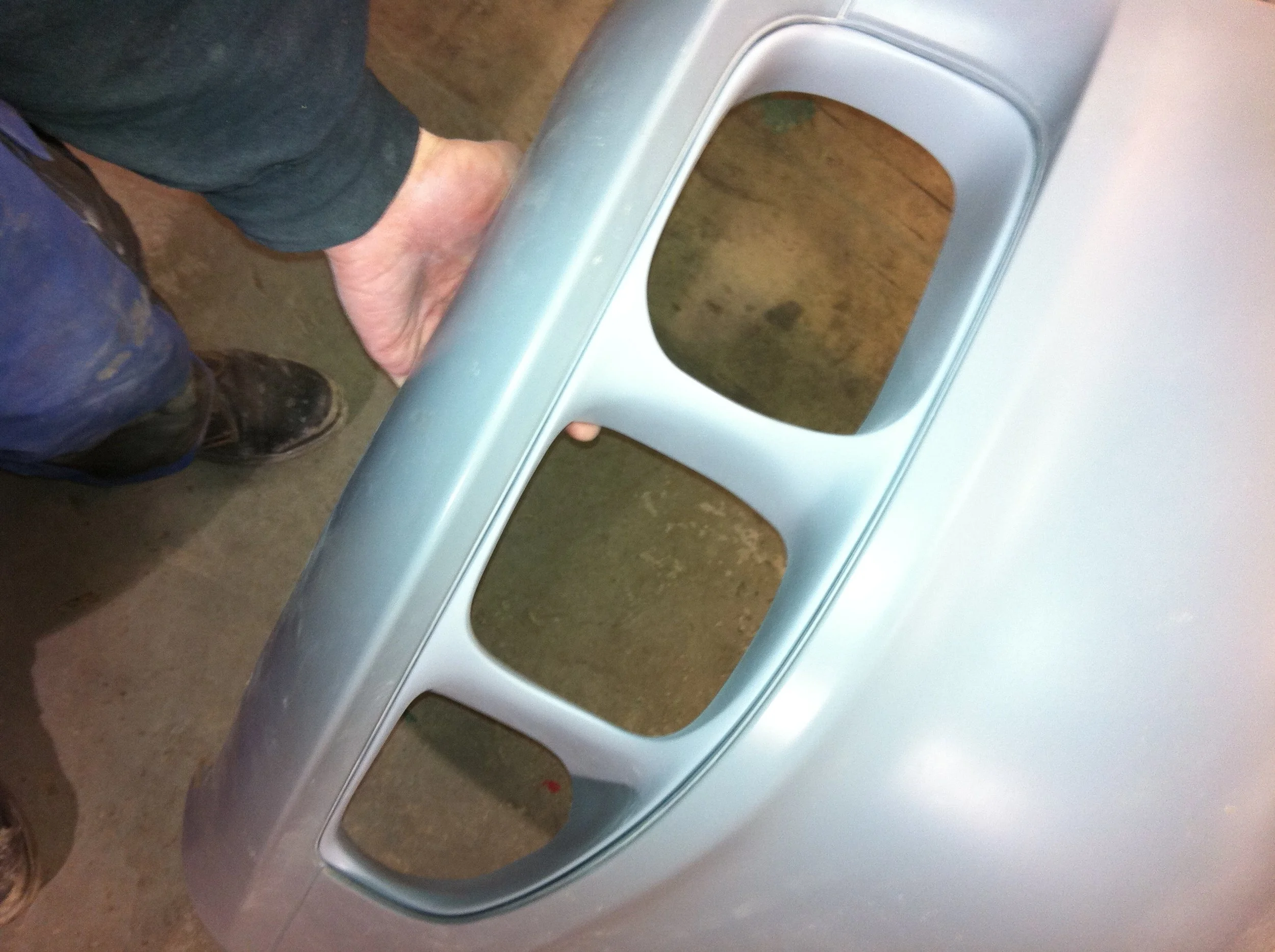 A person holding a silver car bumper with three large cutouts, standing on a concrete floor.