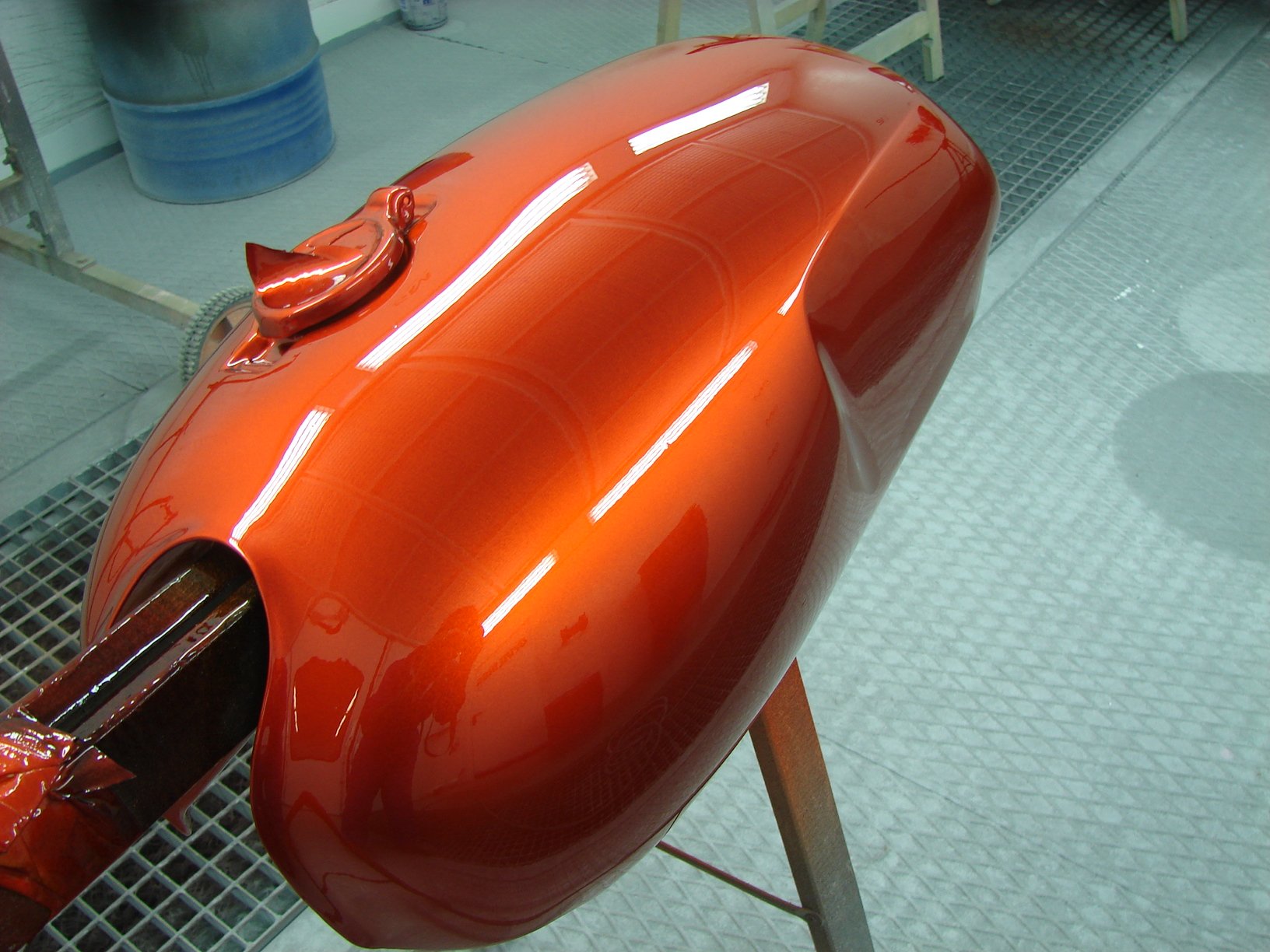 A metallic red-orange motorcycle fuel tank being painted in a spray booth.