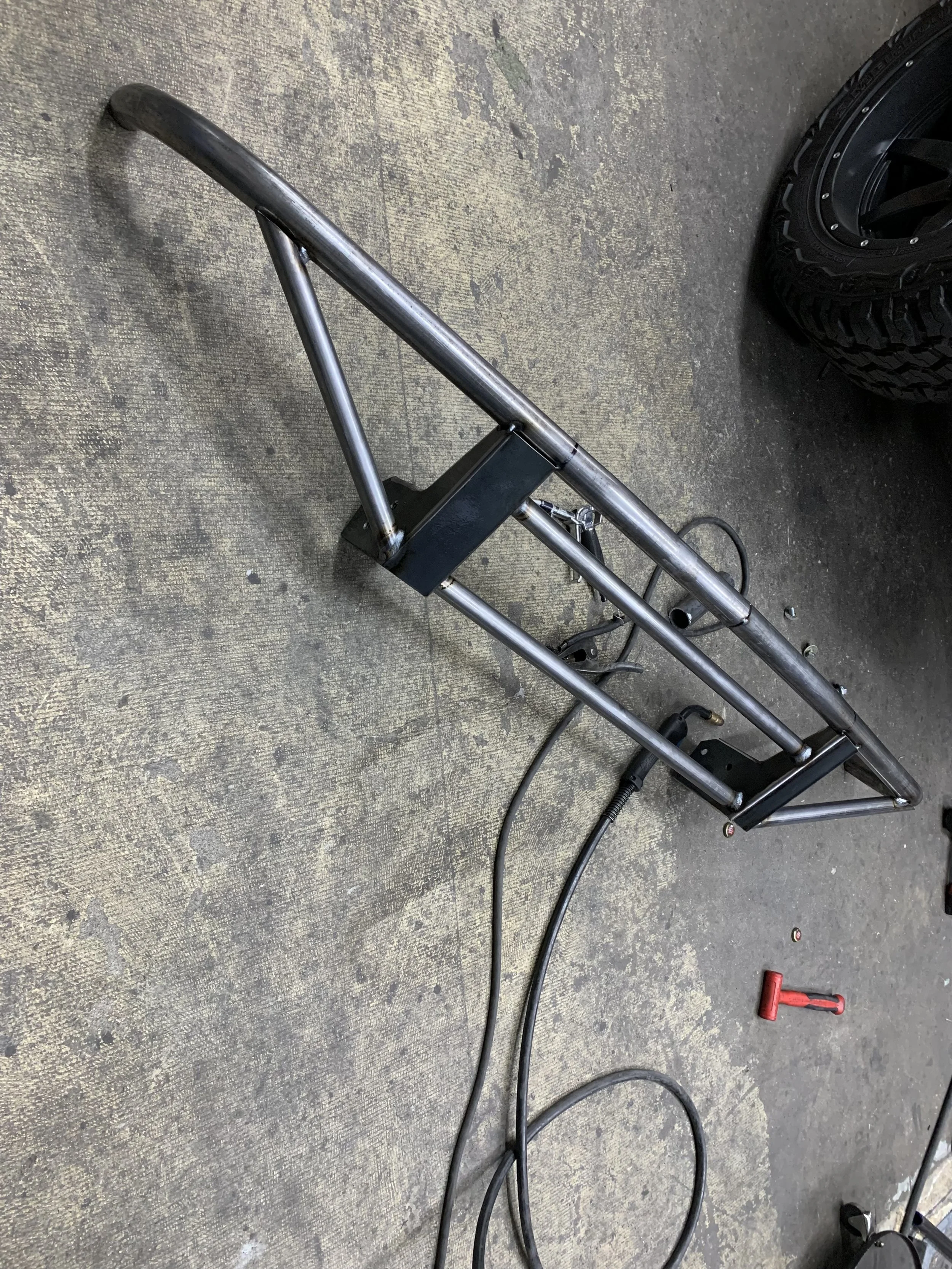Unfinished metal frame, possibly a part of a vehicle or equipment, resting on a concrete floor with a red tool and tire nearby.
